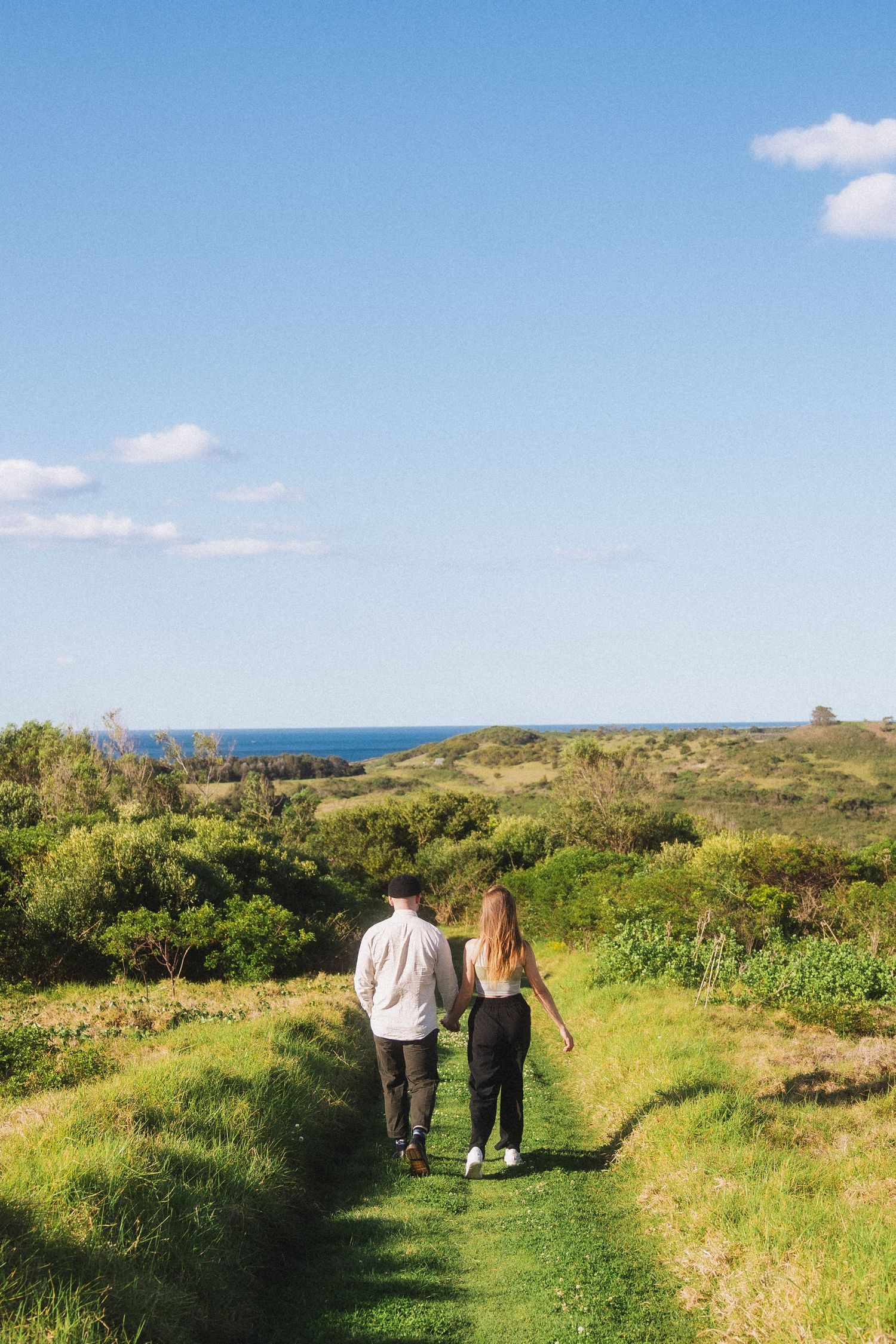 A couple walks along a scenic coastal path with green grass and blue ocean views on a sunny day.