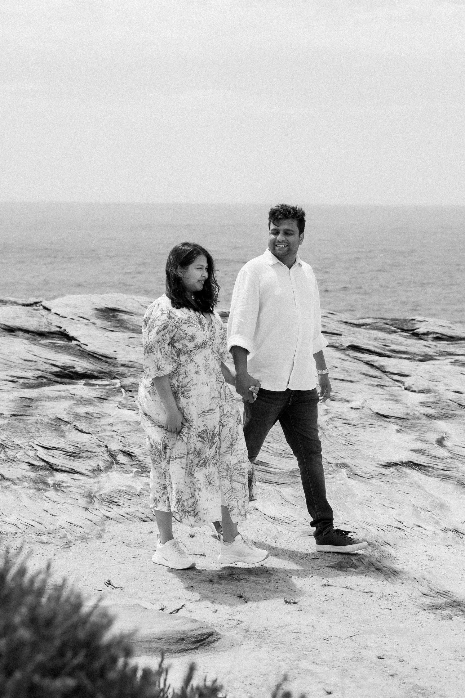A black and white photo captures a tender moment between two people on rocky coastal terrain.