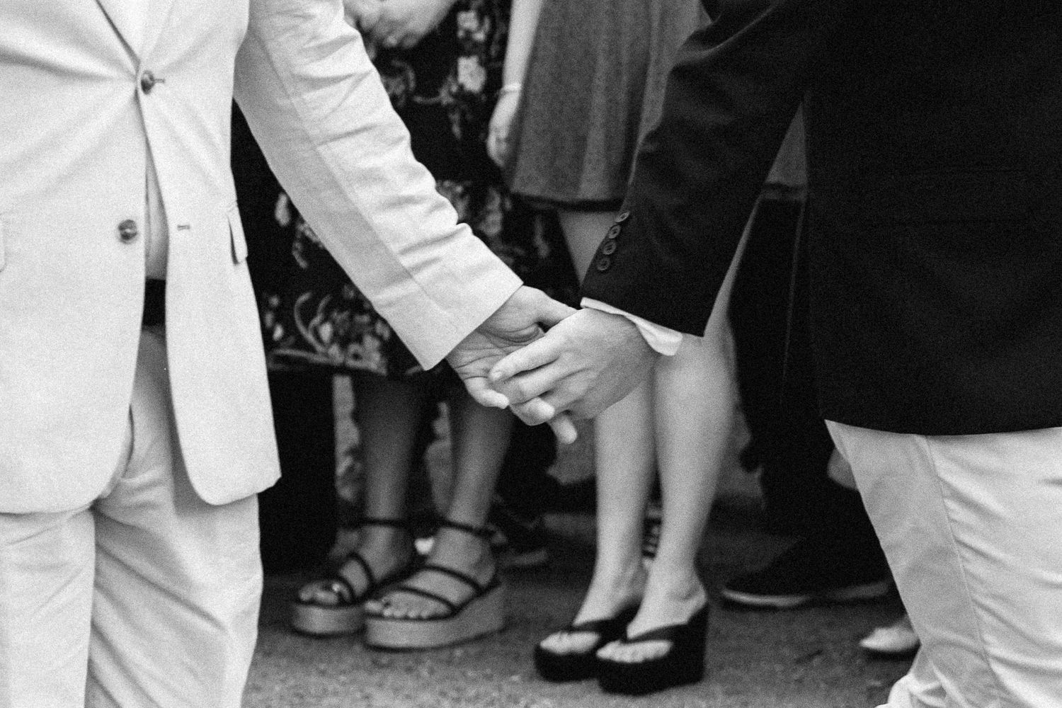 Black and white photo of a couple holding hands at a wedding, wearing formal attire and heels.