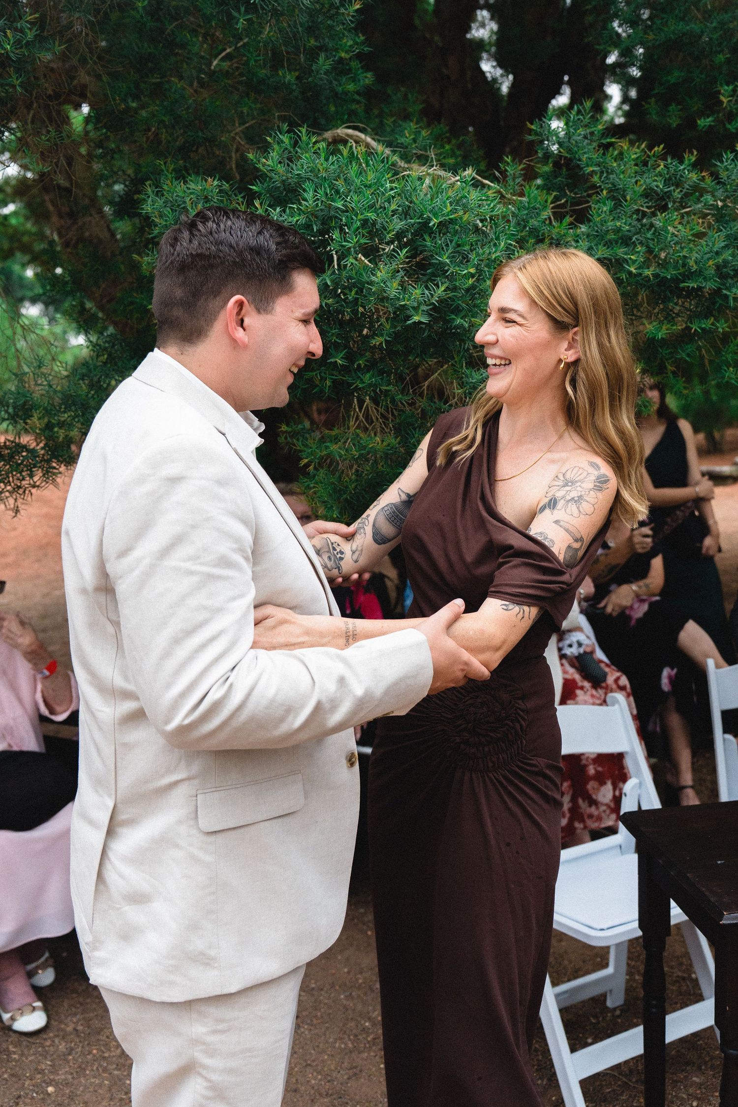 A smiling couple holds hands outdoors at a wedding, man in cream suit, woman in brown dress with tattoos.