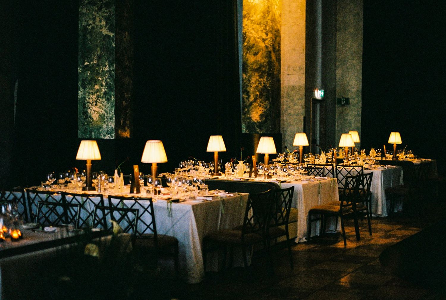 Elegant dimly lit banquet hall with long tables, white linens, glowing lamp centerpieces, and crystal glassware.