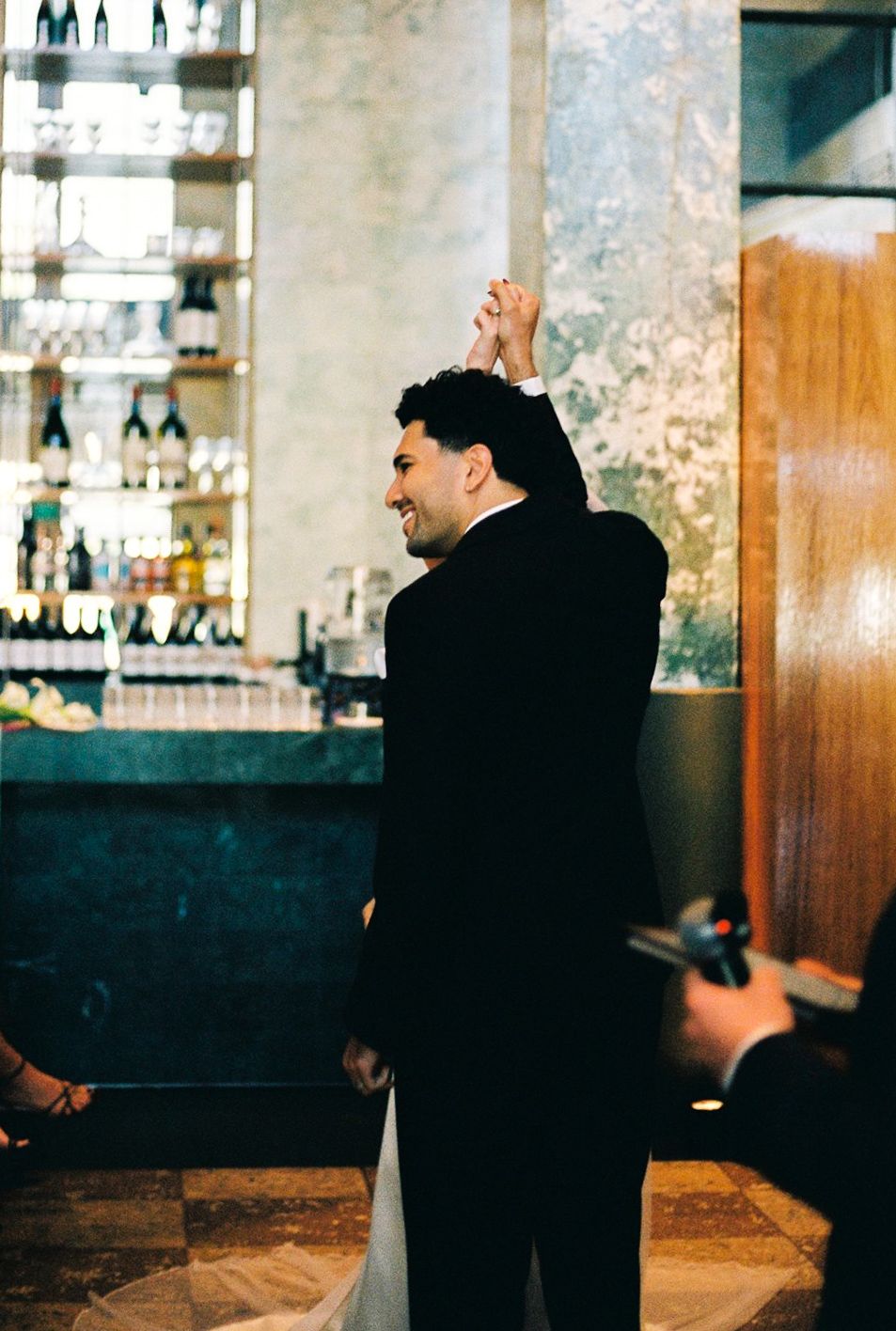 Groom in black tuxedo raises bride's hand at elegant wedding reception with bar backdrop.