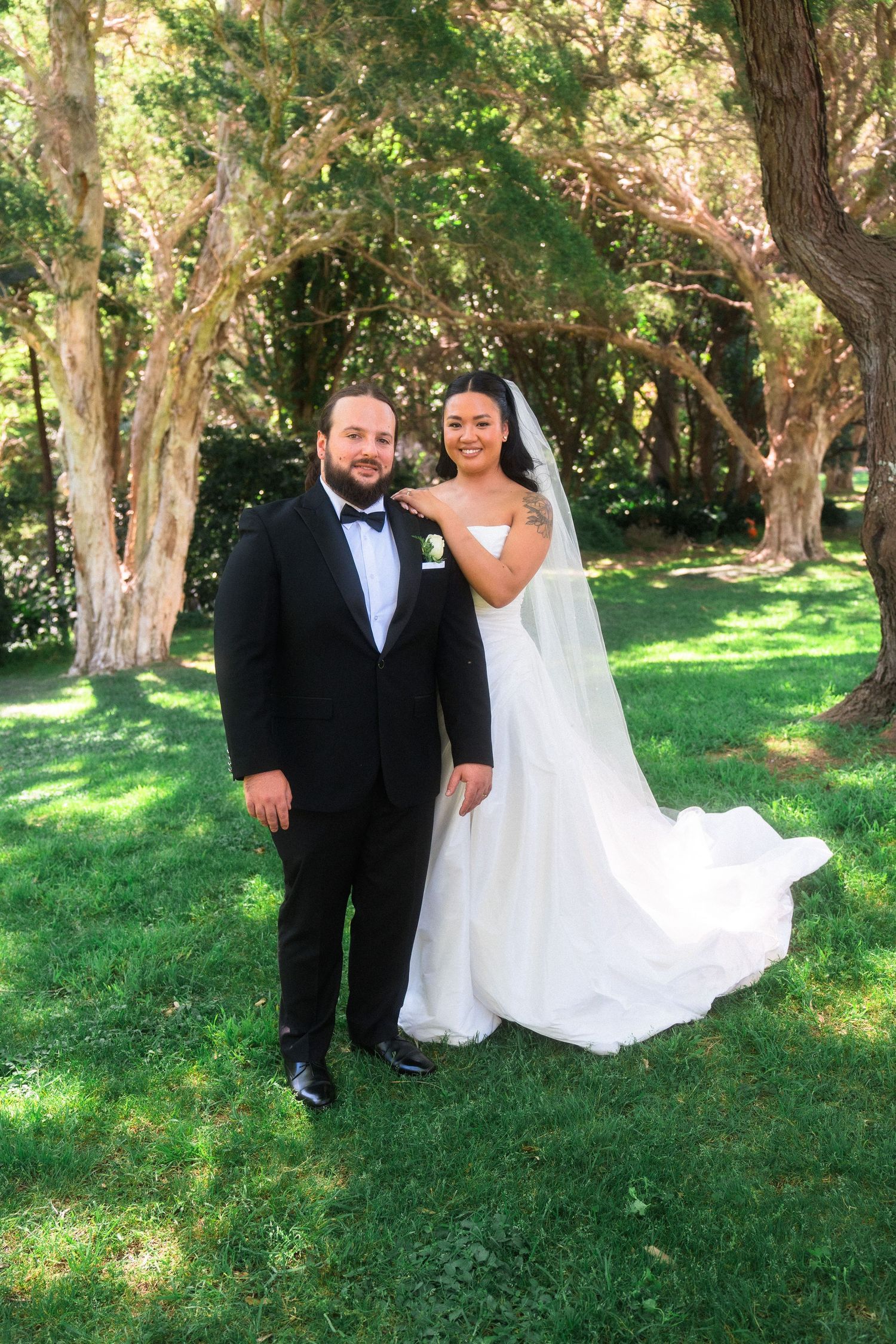 Newlyweds pose together on green grass with trees in the background.