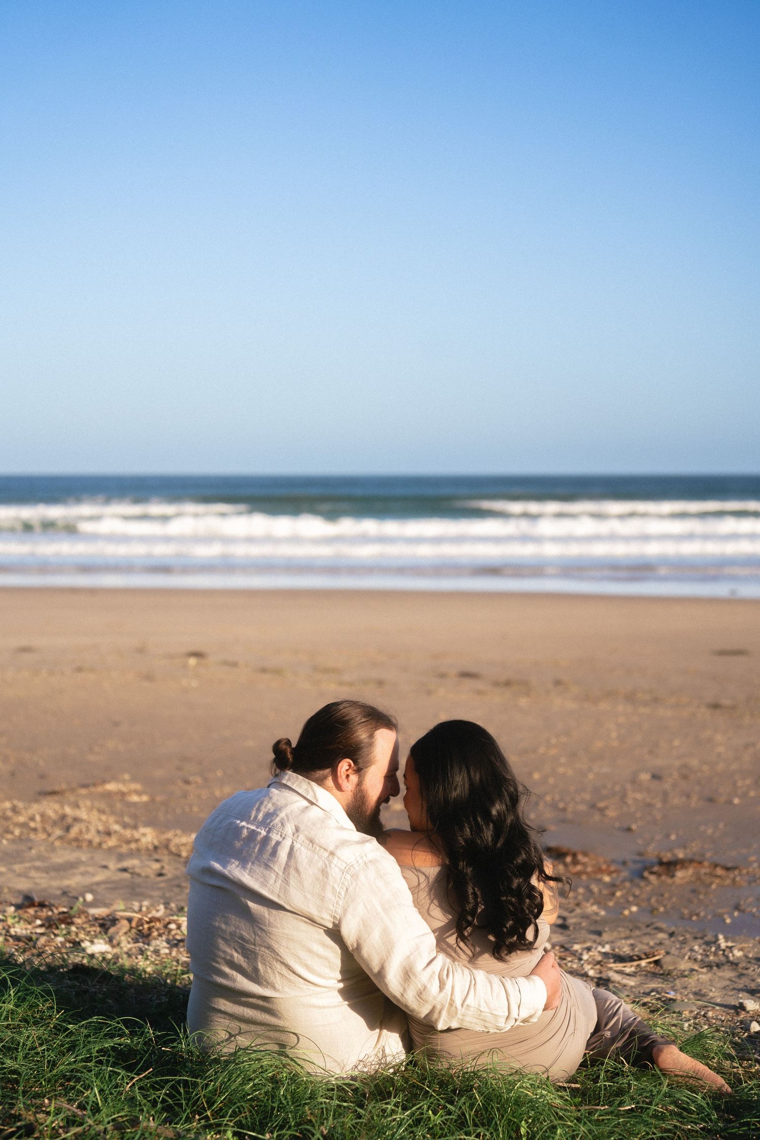 A couple embraces while sitting on a beach overlooking the ocean.