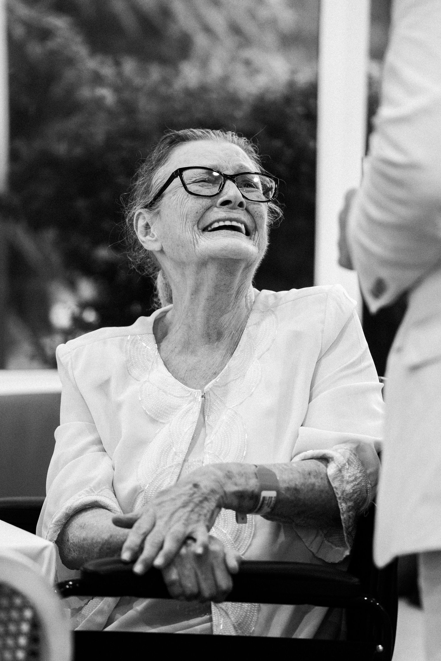 Elderly woman with glasses laughing joyfully while shaking hands, black and white portrait photo.