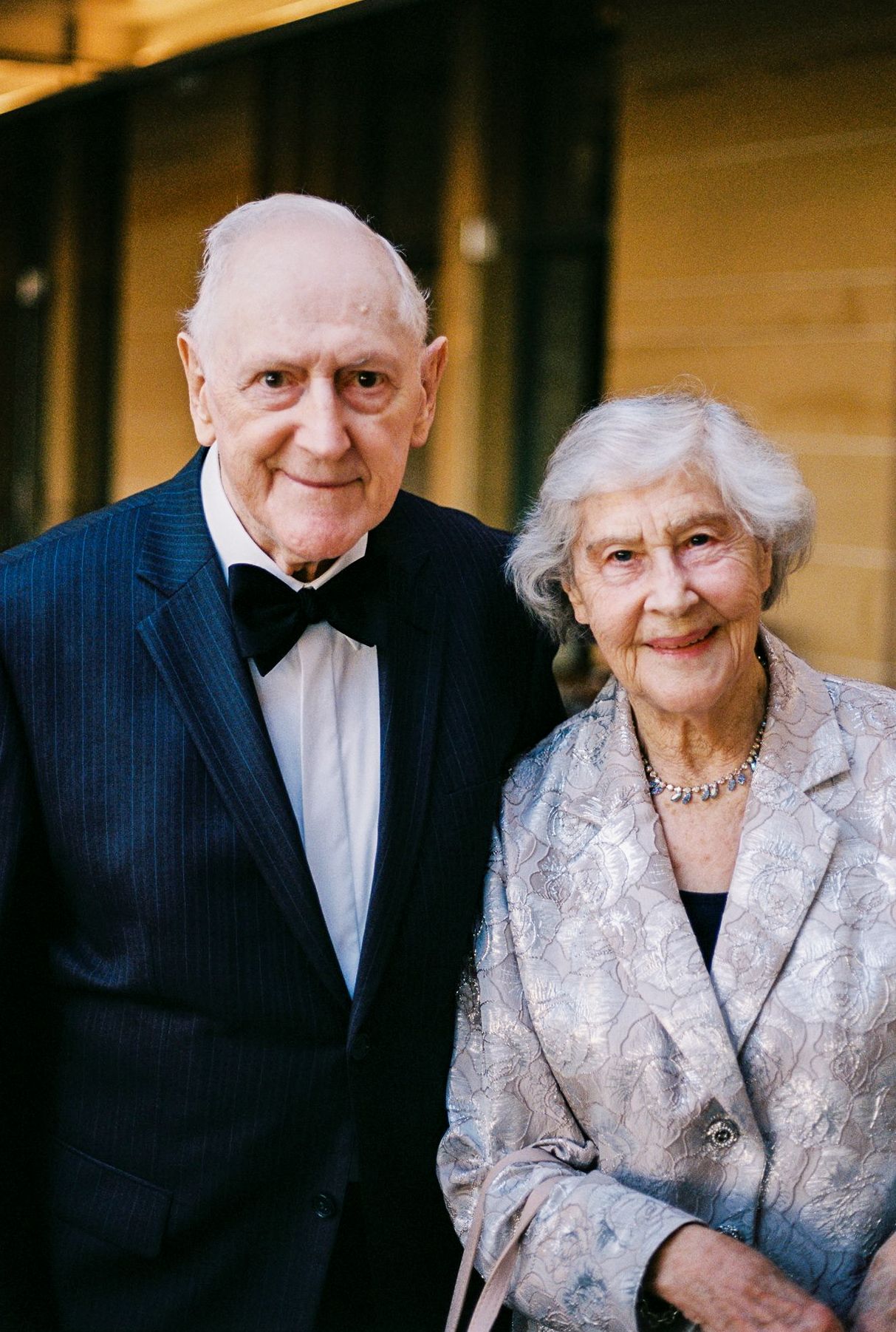 Elderly couple dressed formally, man in tuxedo and woman in silver blazer, smiling together outdoors.