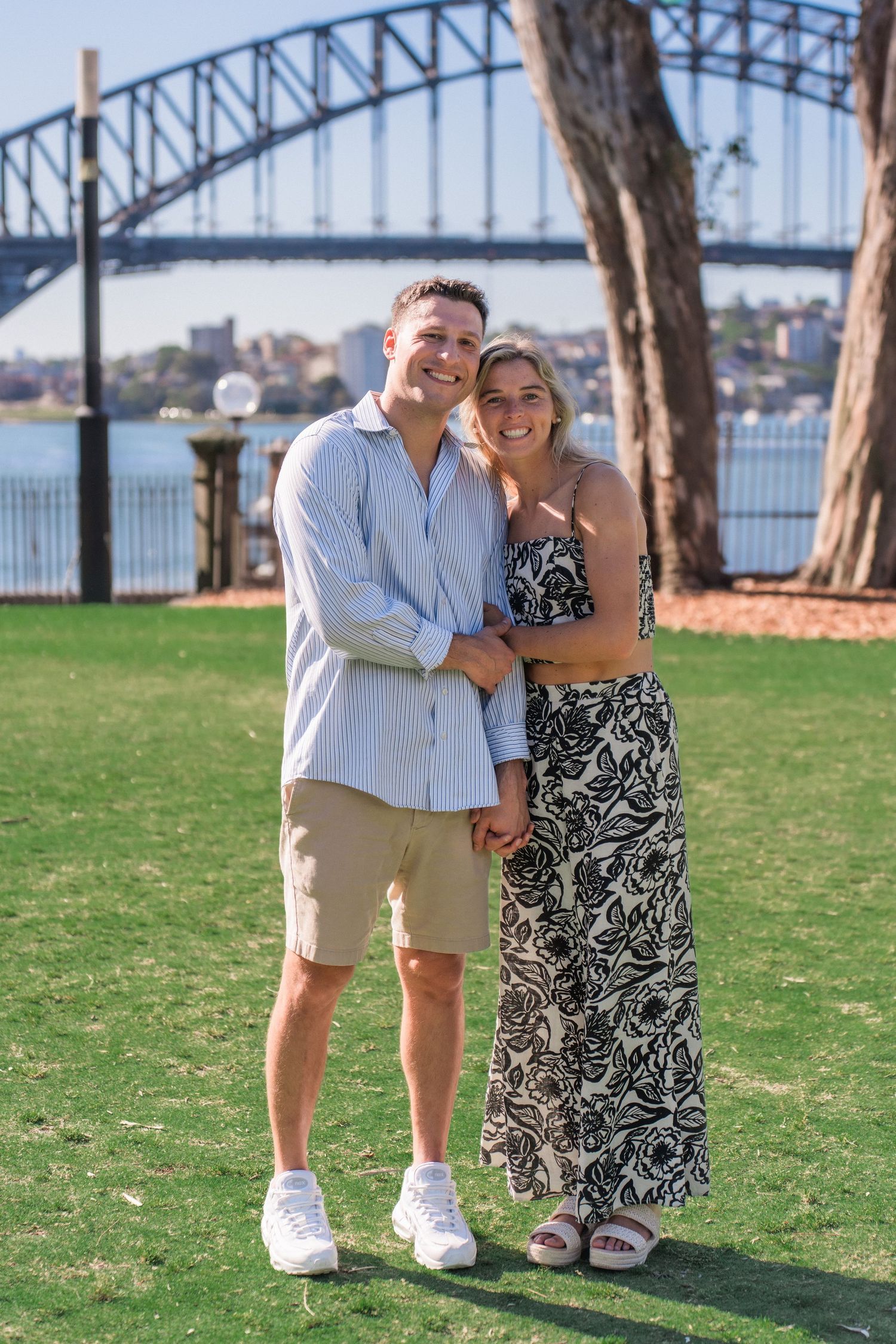 A couple poses together on a grassy lawn with the Sydney Harbour Bridge visible in the background.