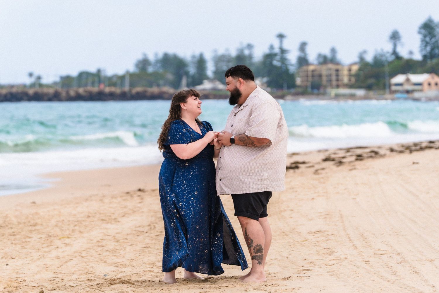 A couple embraces on a sandy beach with ocean waves in the background.