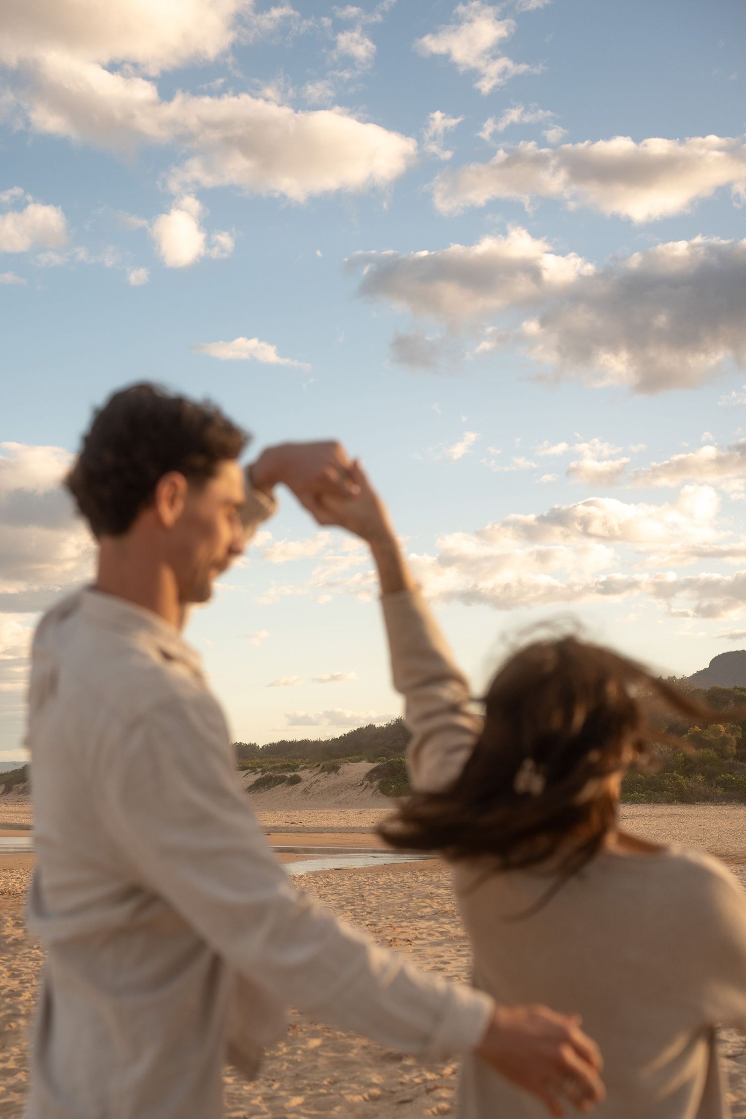 Playful couple dancing and spinning on beach with cloudy sky backdrop.