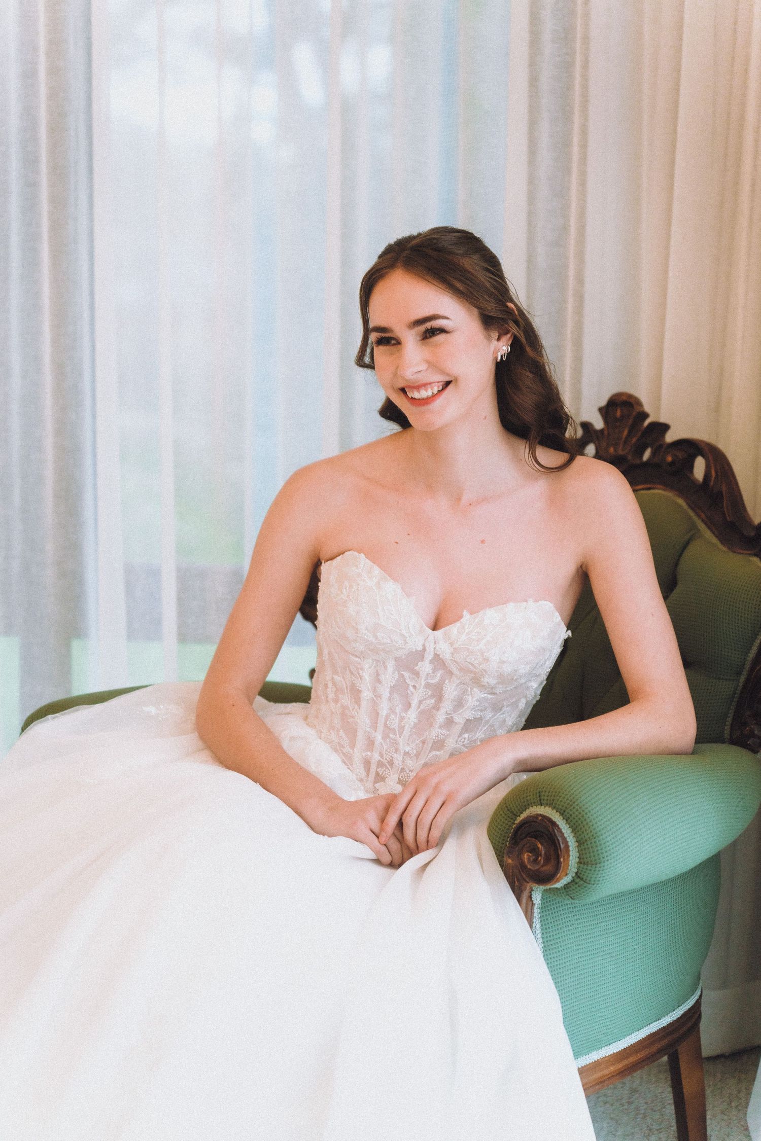Bride in strapless white lace wedding gown poses elegantly on mint green chair in natural light.