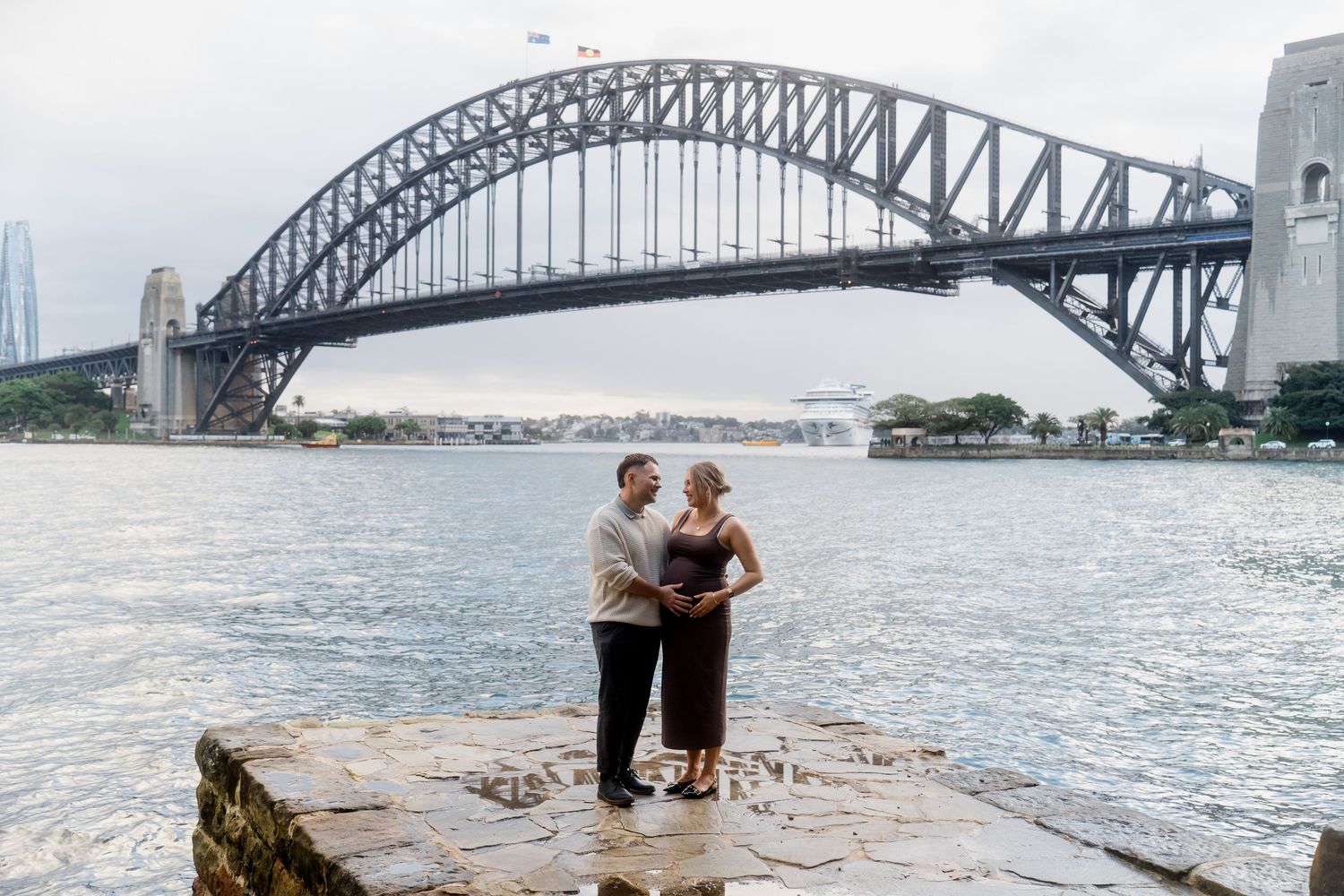 A couple embraces while standing on rocks with the Sydney Harbour Bridge in the background.