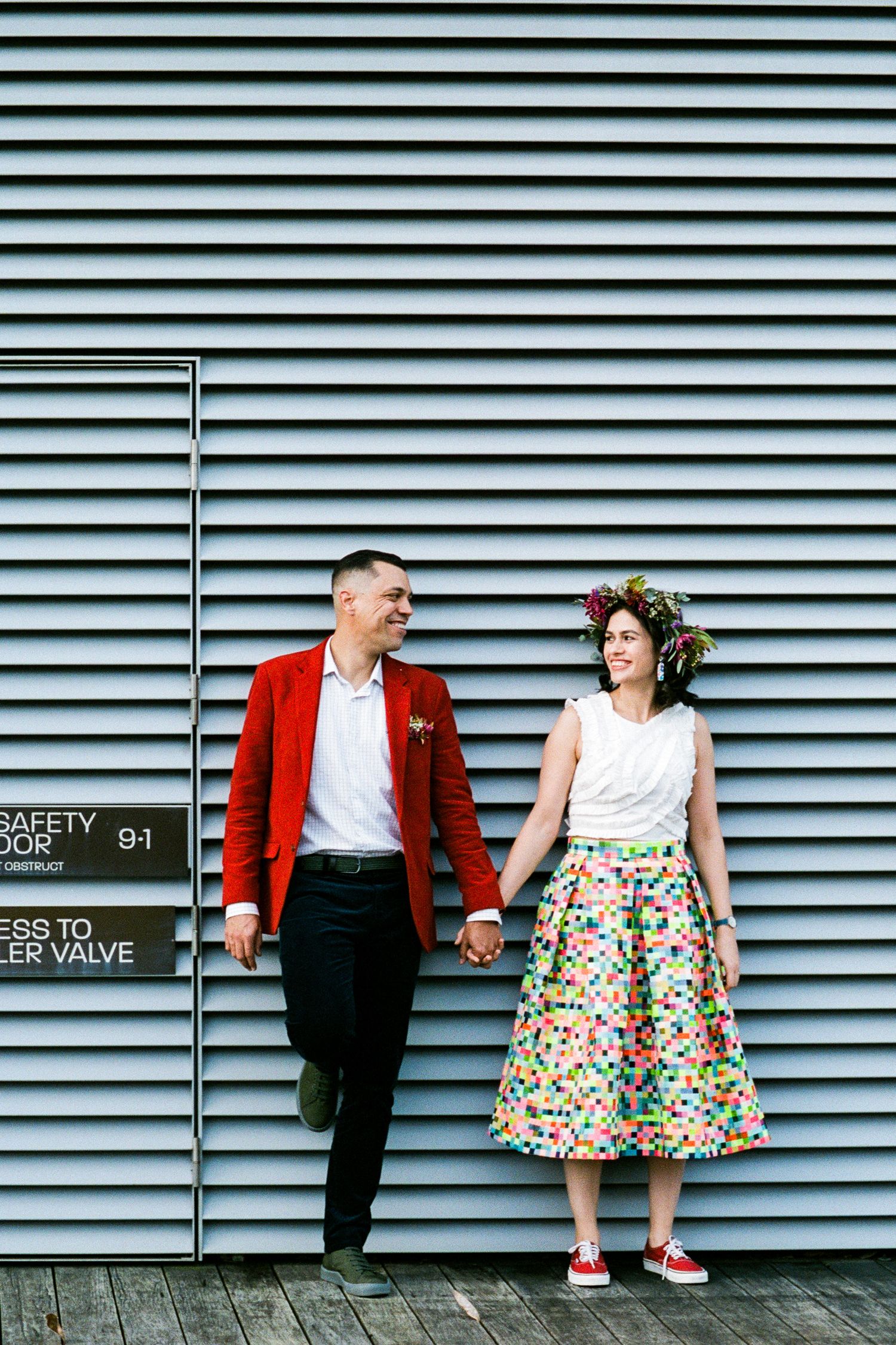 Stylish couple in red jacket and floral skirt posing against industrial metal wall.