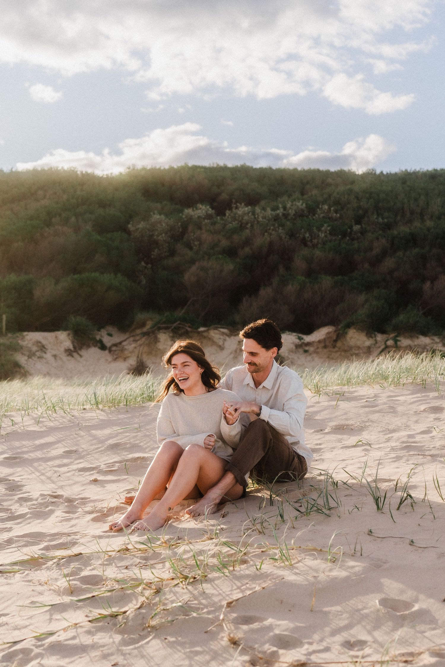 Couple relaxes together on sandy dunes with coastal vegetation in background.