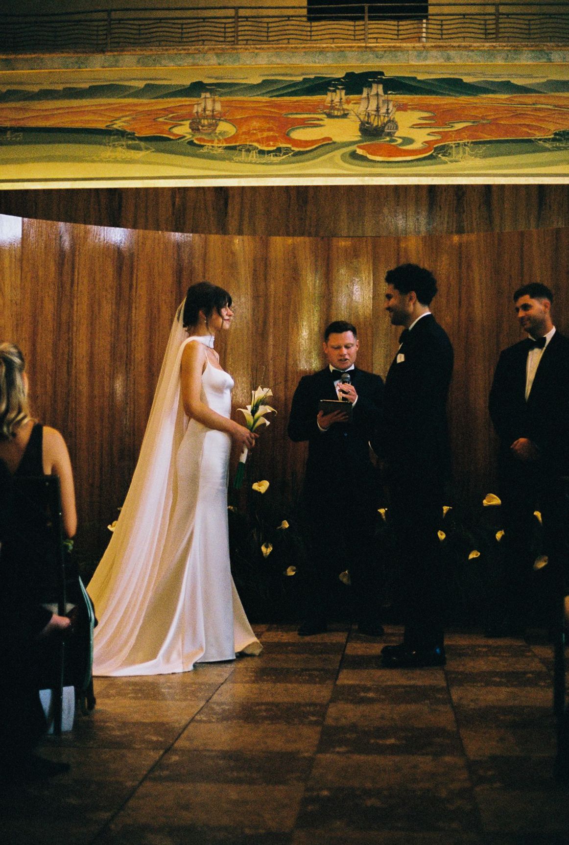 Bride and groom exchange vows at elegant indoor wedding ceremony with wood-paneled walls and decorative mural backdrop.