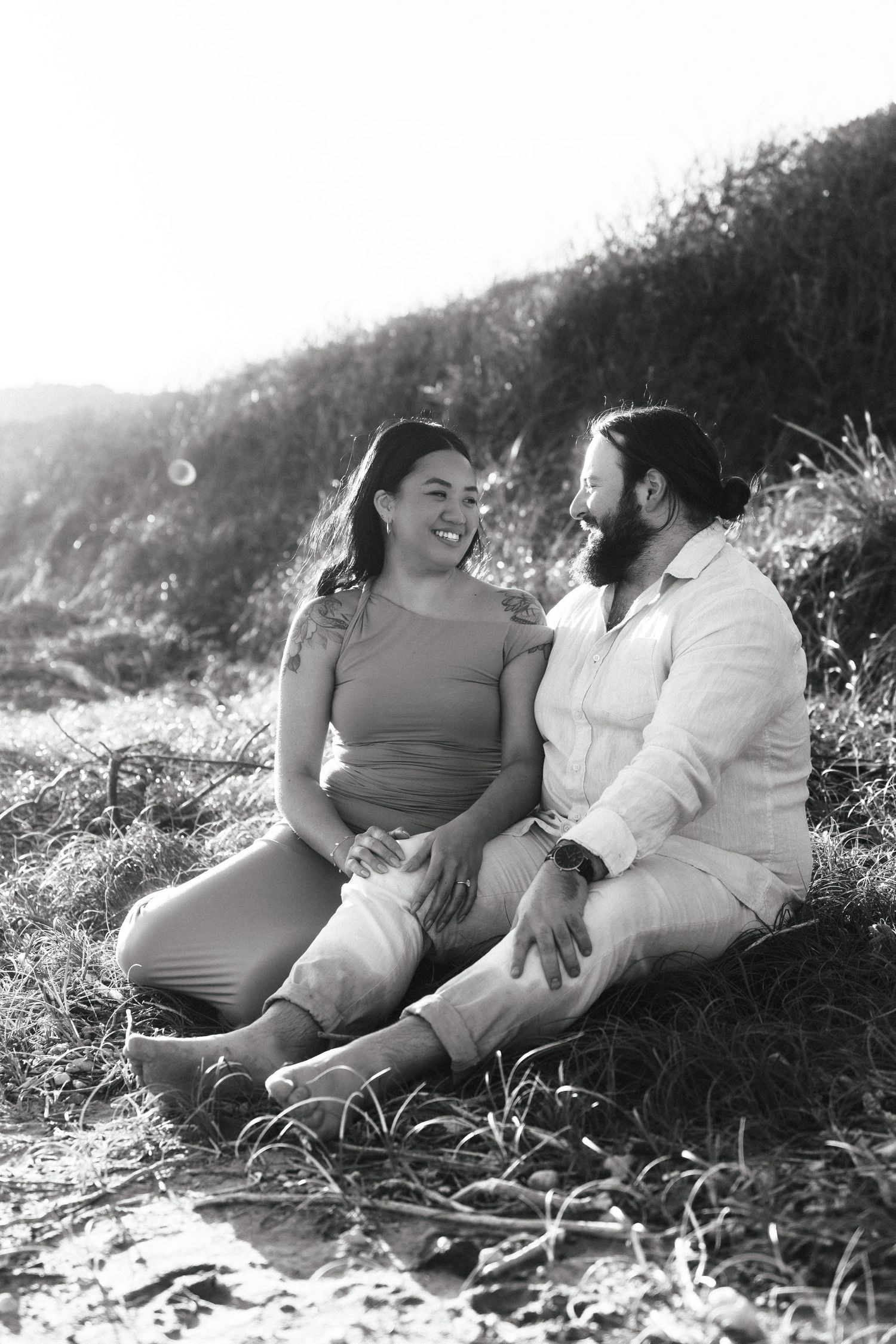 A couple sits together on a grassy hillside sharing a candid moment.