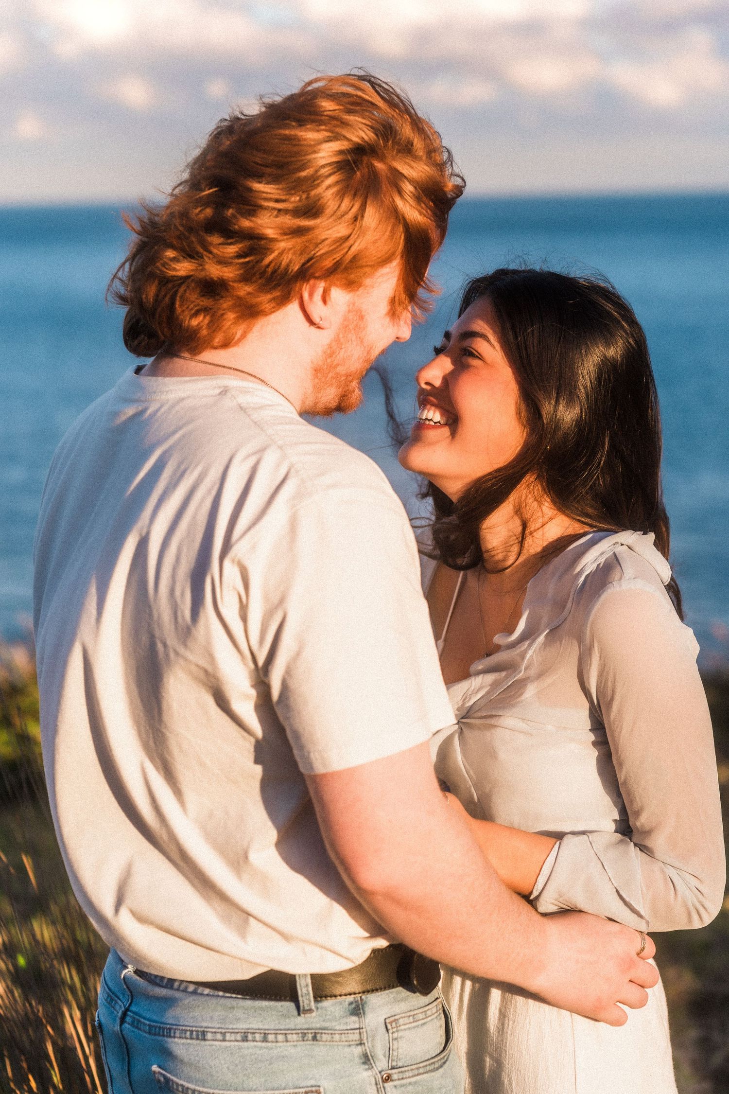 A romantic couple embraces against a coastal sunset backdrop.