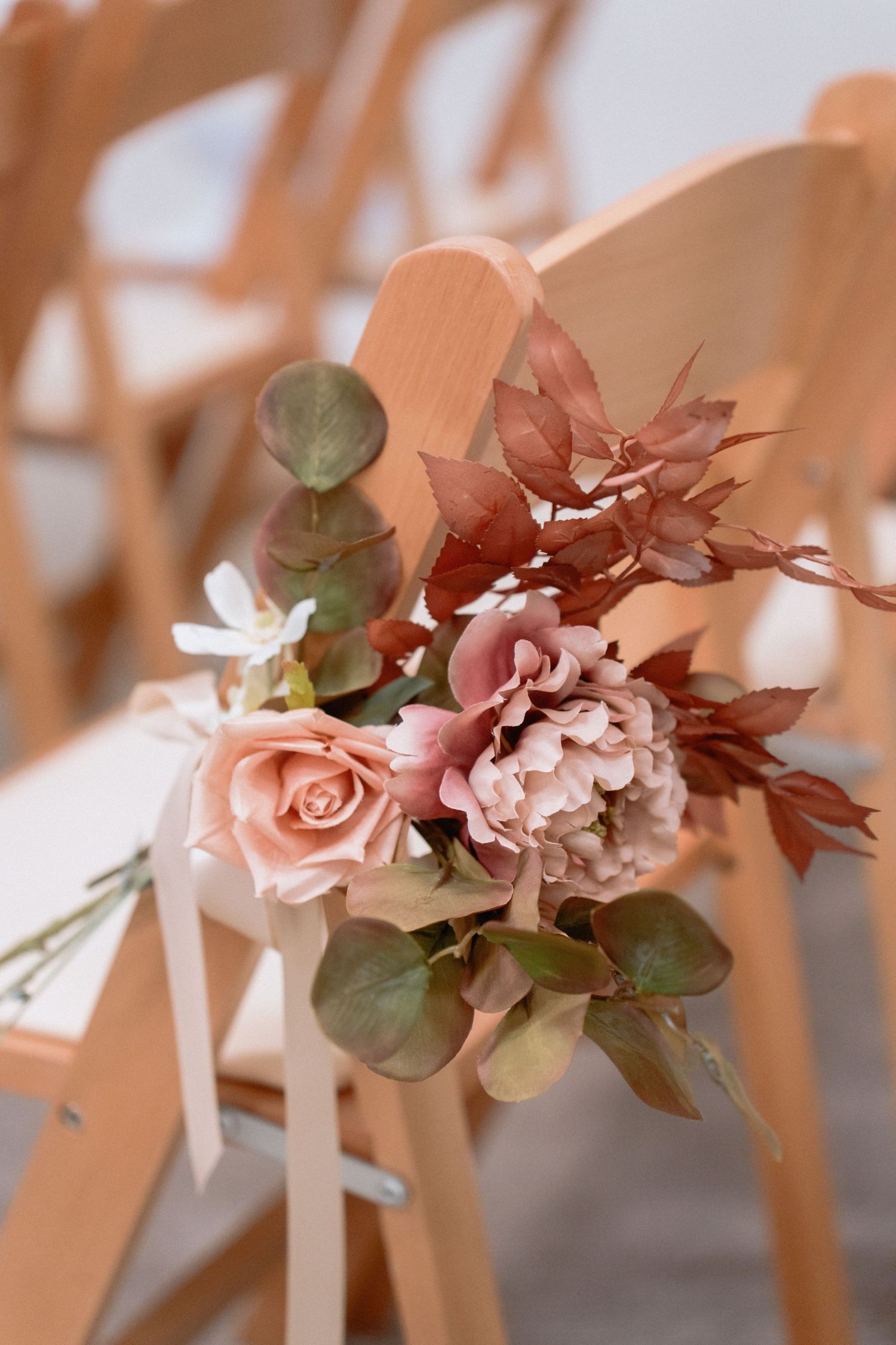 Romantic blush pink floral decorations adorn wooden chairs for a wedding ceremony.