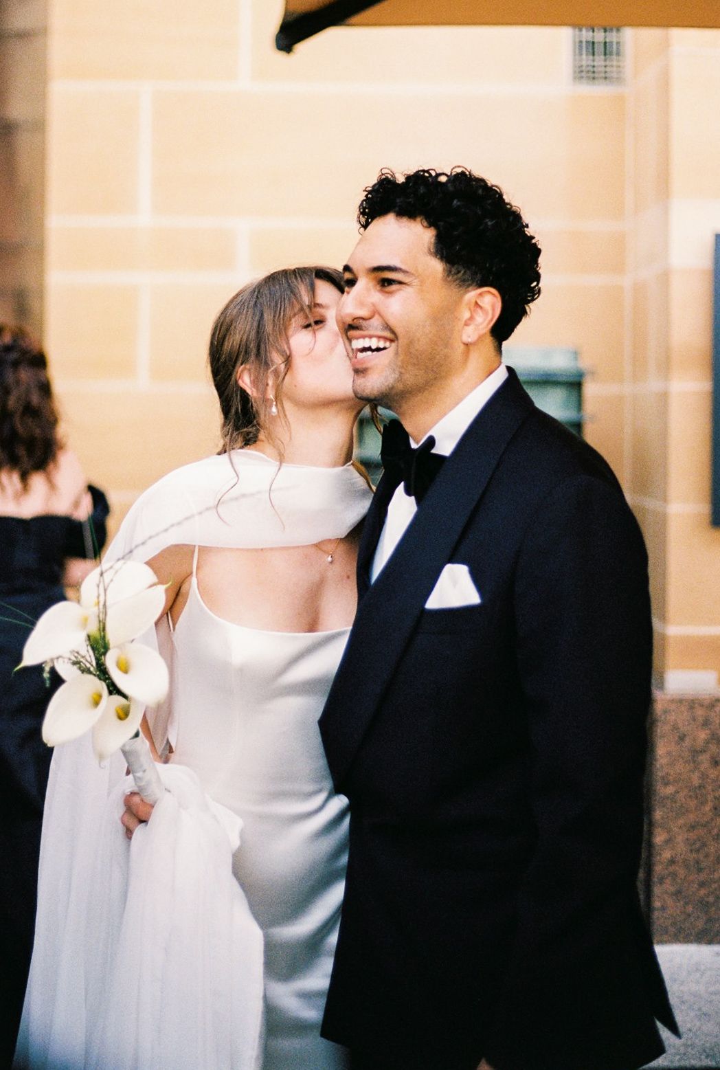 Bride in white gown kisses groom in black tuxedo holding calla lilies at elegant outdoor wedding ceremony.