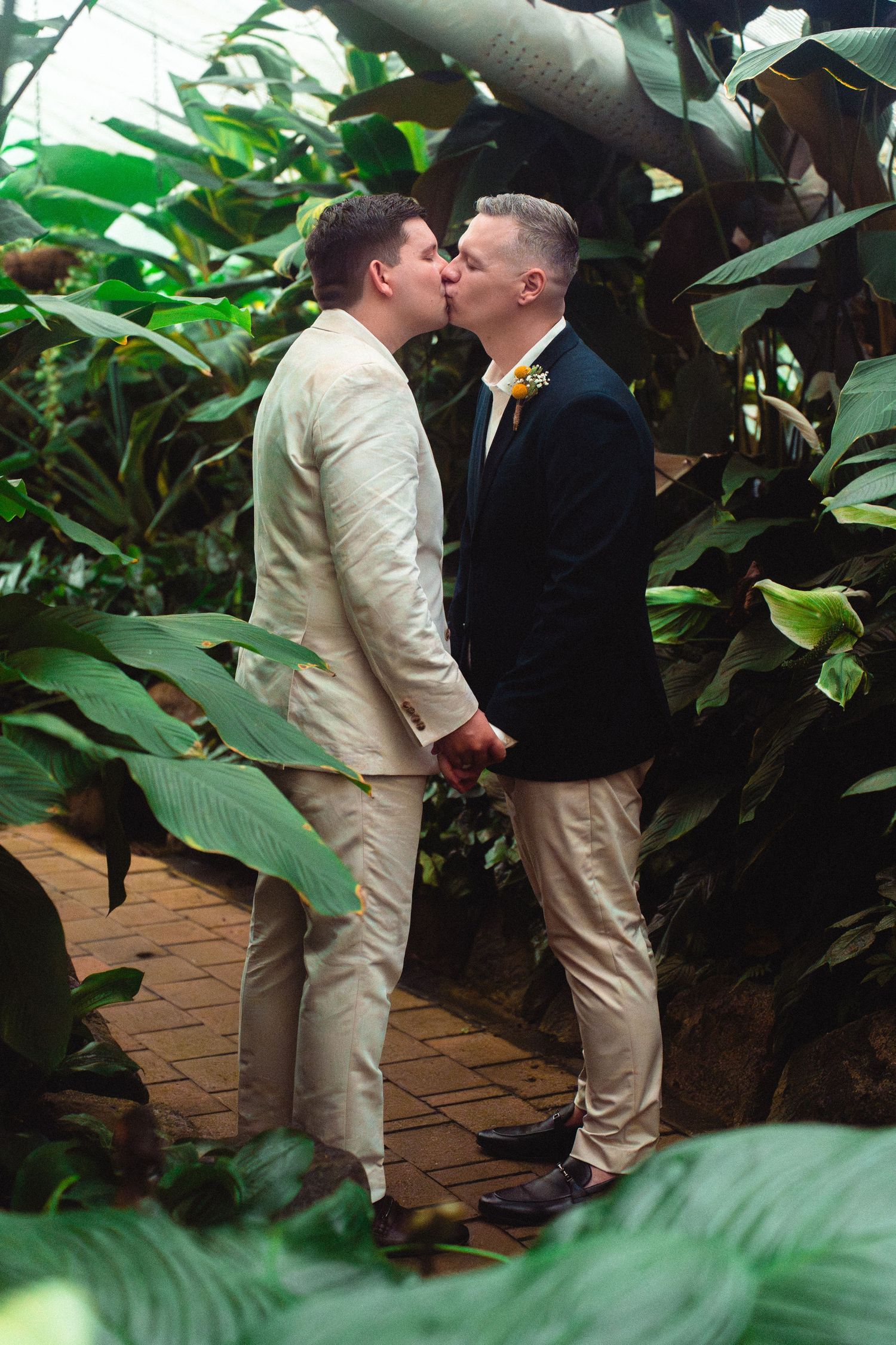 Two grooms share a kiss in a lush tropical greenhouse setting on their wedding day.