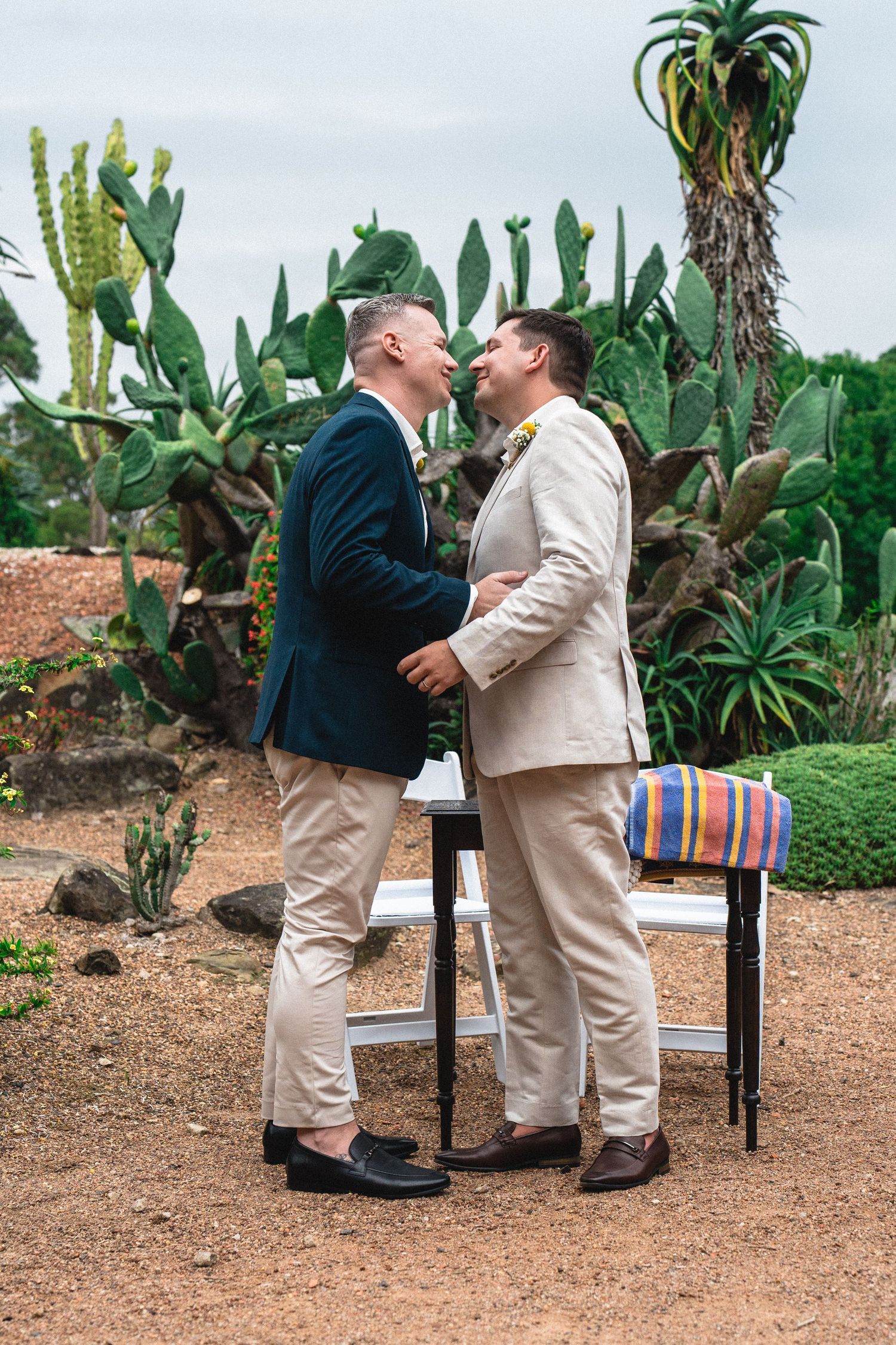 Two grooms share a joyful moment at an outdoor wedding ceremony surrounded by cacti and tropical plants.
