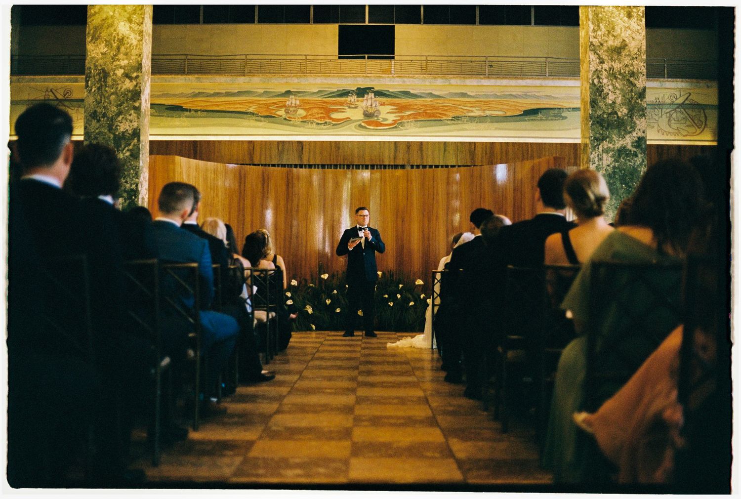 Elegant event speaker addresses seated audience in ornate wood-paneled hall with decorative murals and warm lighting.