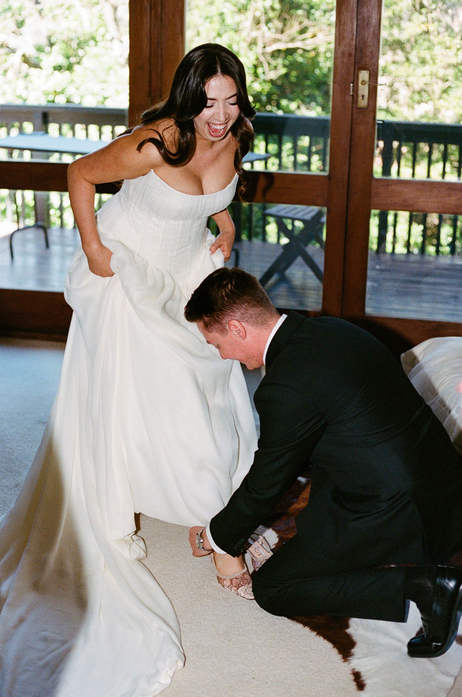 Groom helping bride with her wedding shoes on a wooden porch.