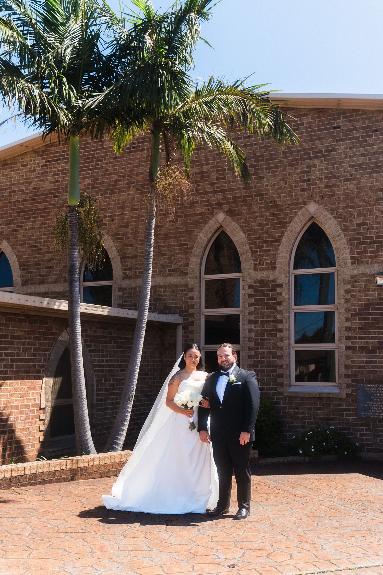 Wedding couple poses outside brick church building with palm trees.