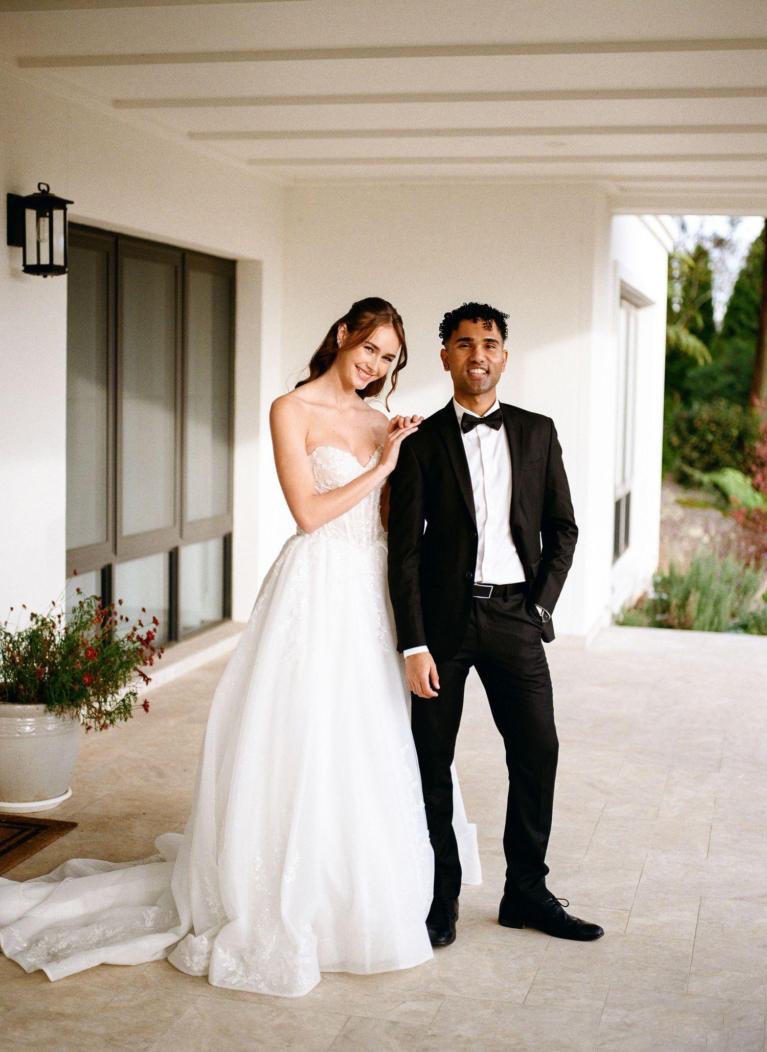 A couple in wedding attire walks together along a bright white columned porch.
