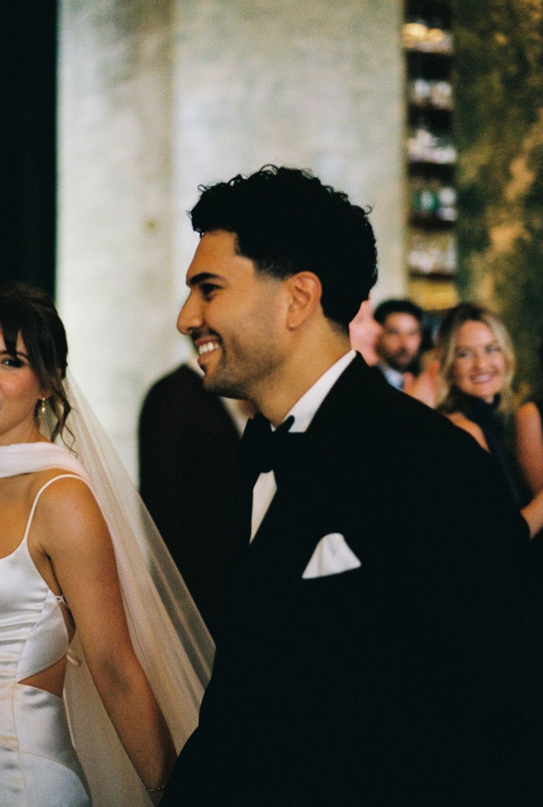 Groom in black tuxedo smiling during wedding ceremony with bride in white gown and veil beside him.