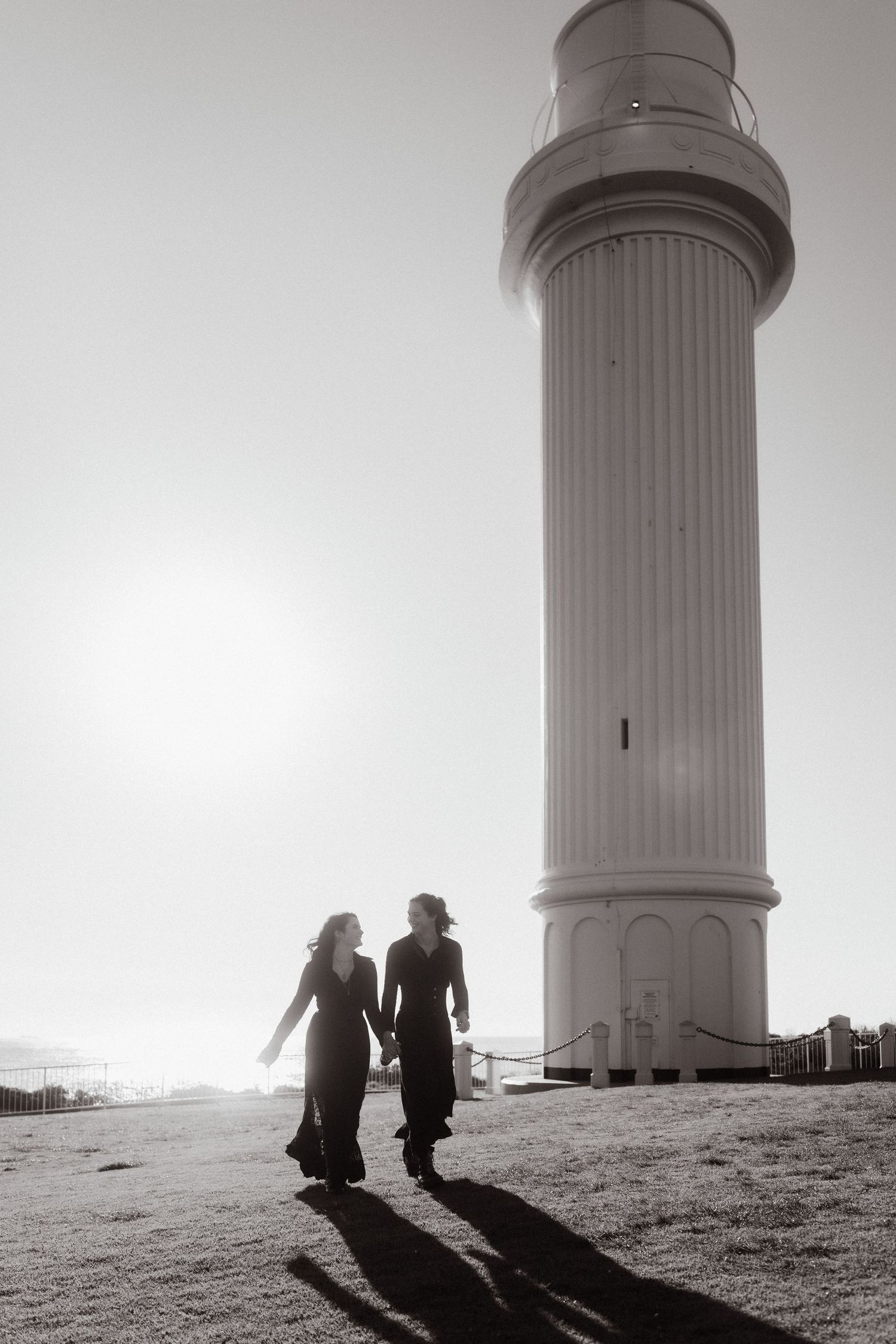 Two figures cast long shadows while standing near a tall white lighthouse structure.