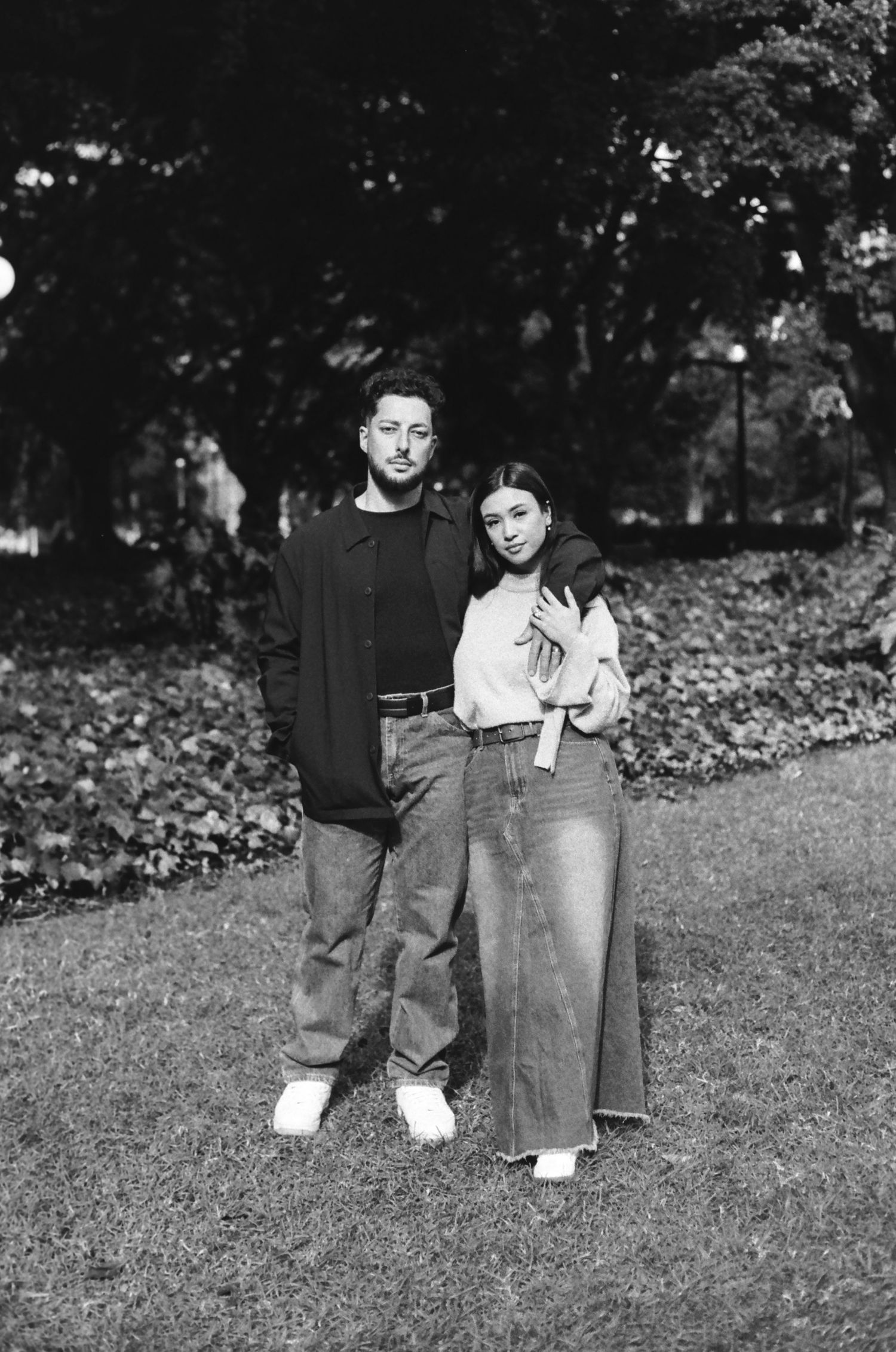 A couple in casual wear stands together on grass with trees in the background.