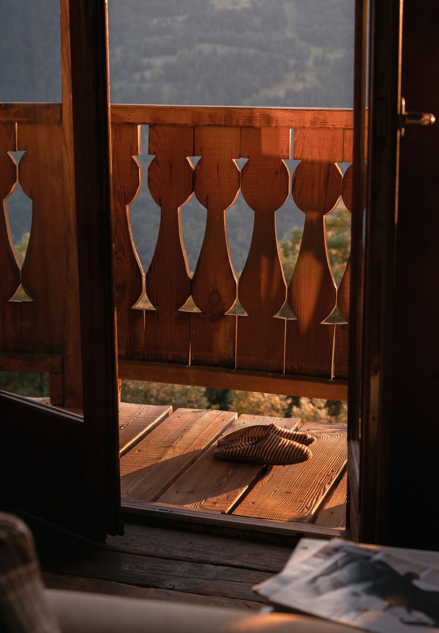 Warm golden sunset light illuminates a wooden chalet balcony with decorative railings and woven sandals on the floor.