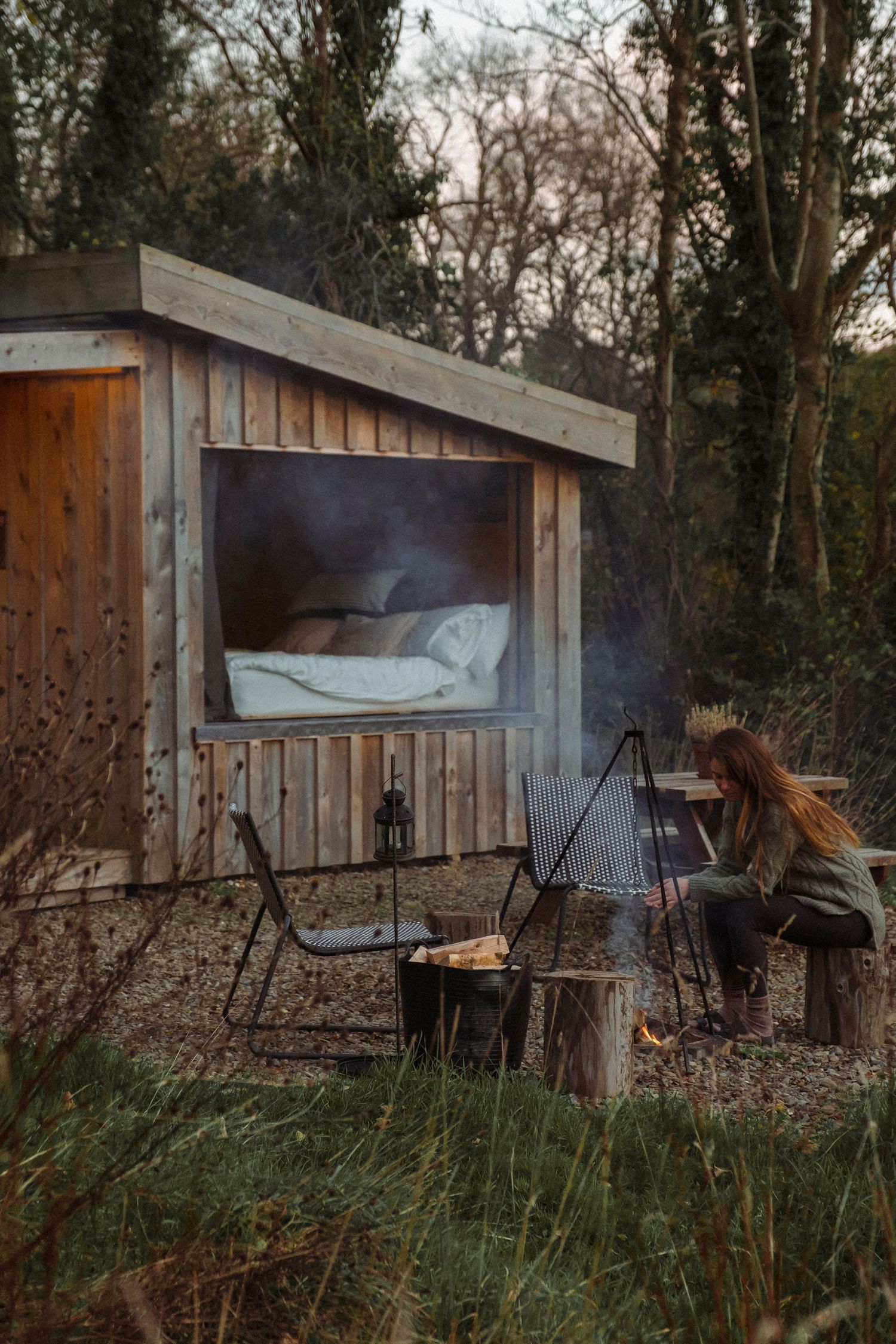 Cozy wooden cabin shelter with outdoor seating area and campfire in misty forest setting.