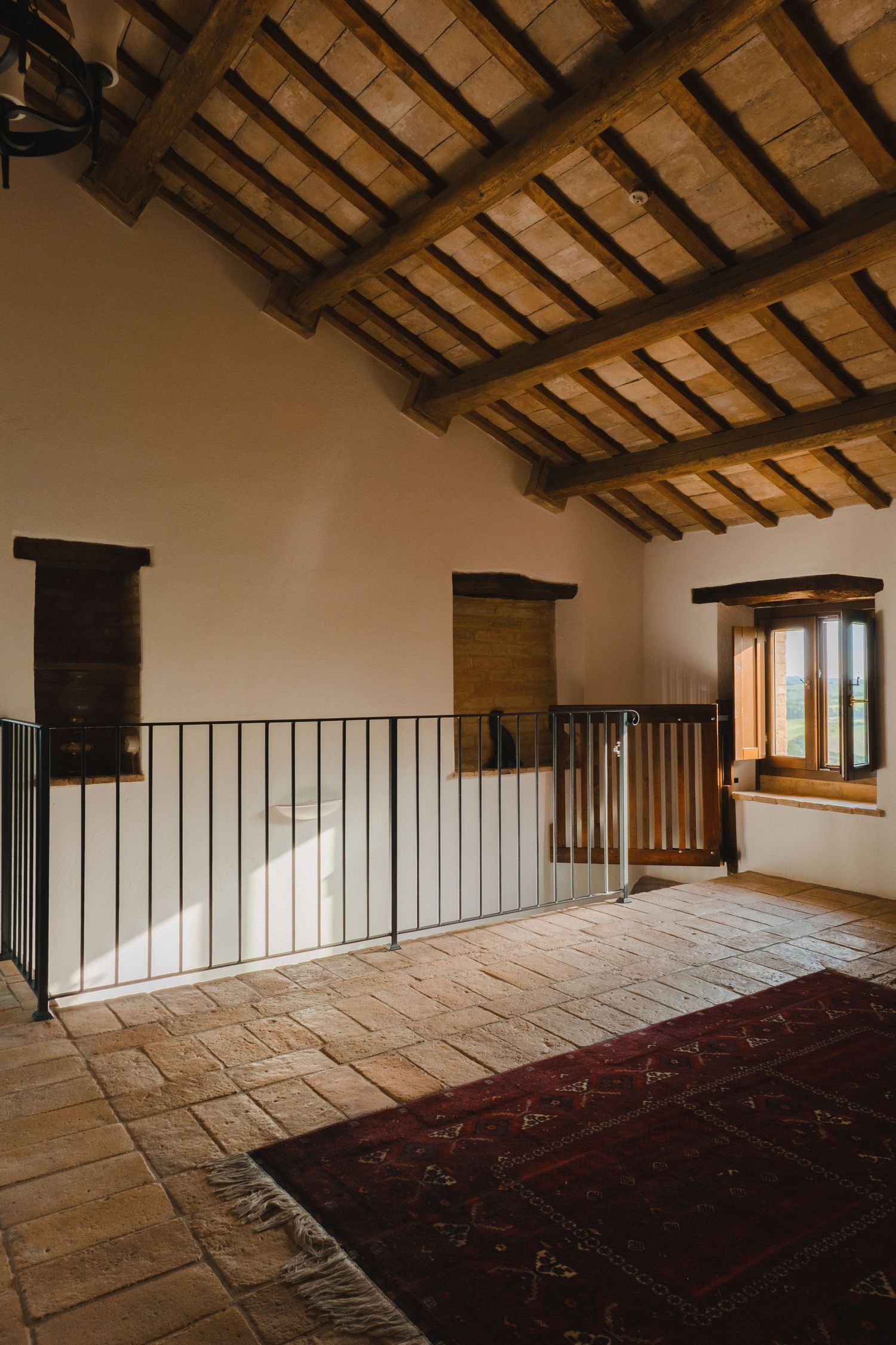Rustic Italian villa upper floor with exposed wooden beam ceiling, stone tile floor, red rug, and wrought iron railing.