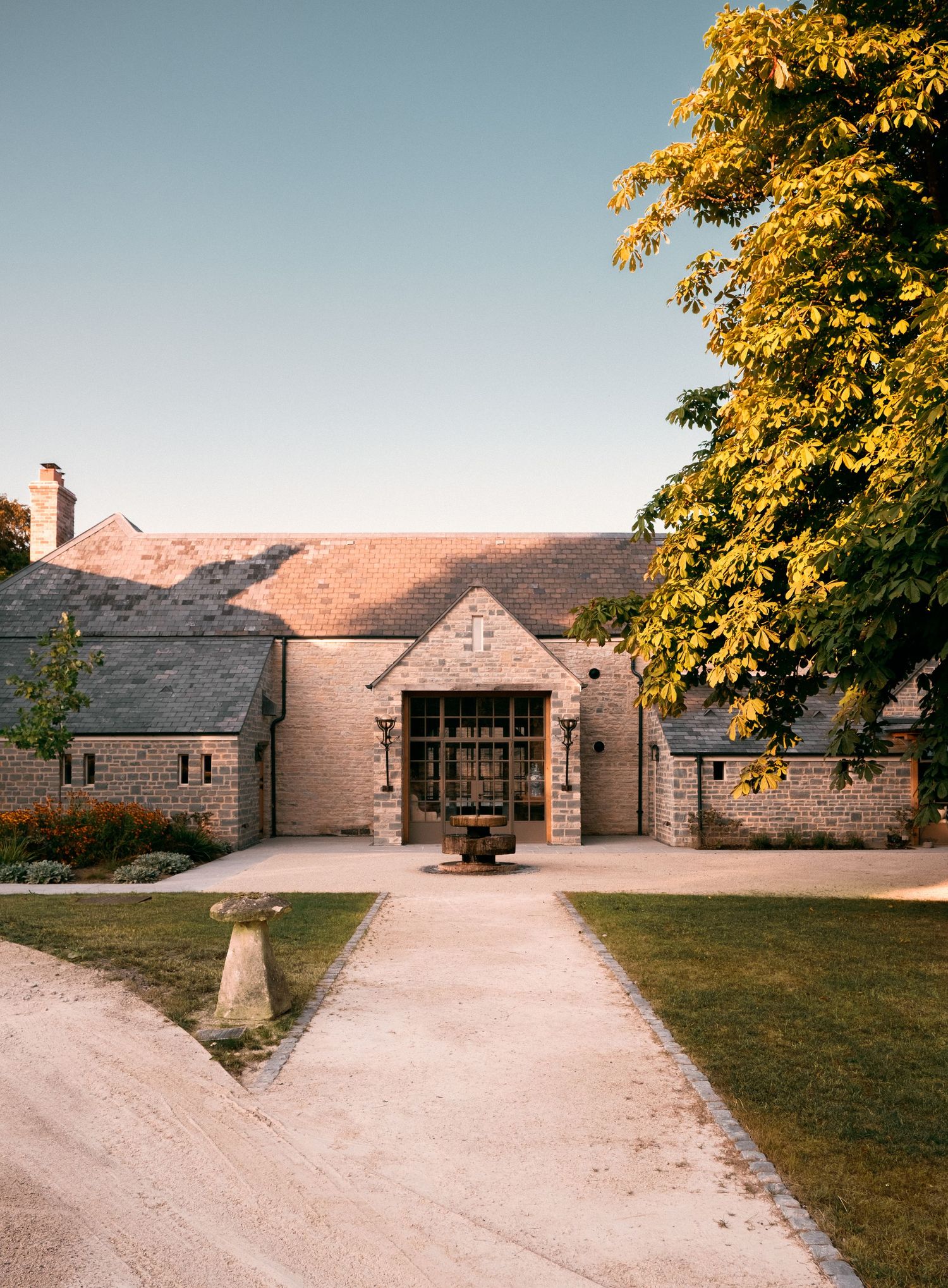 Stone country estate with arched entrance, gravel pathway, manicured lawn, and golden tree under clear blue sky.