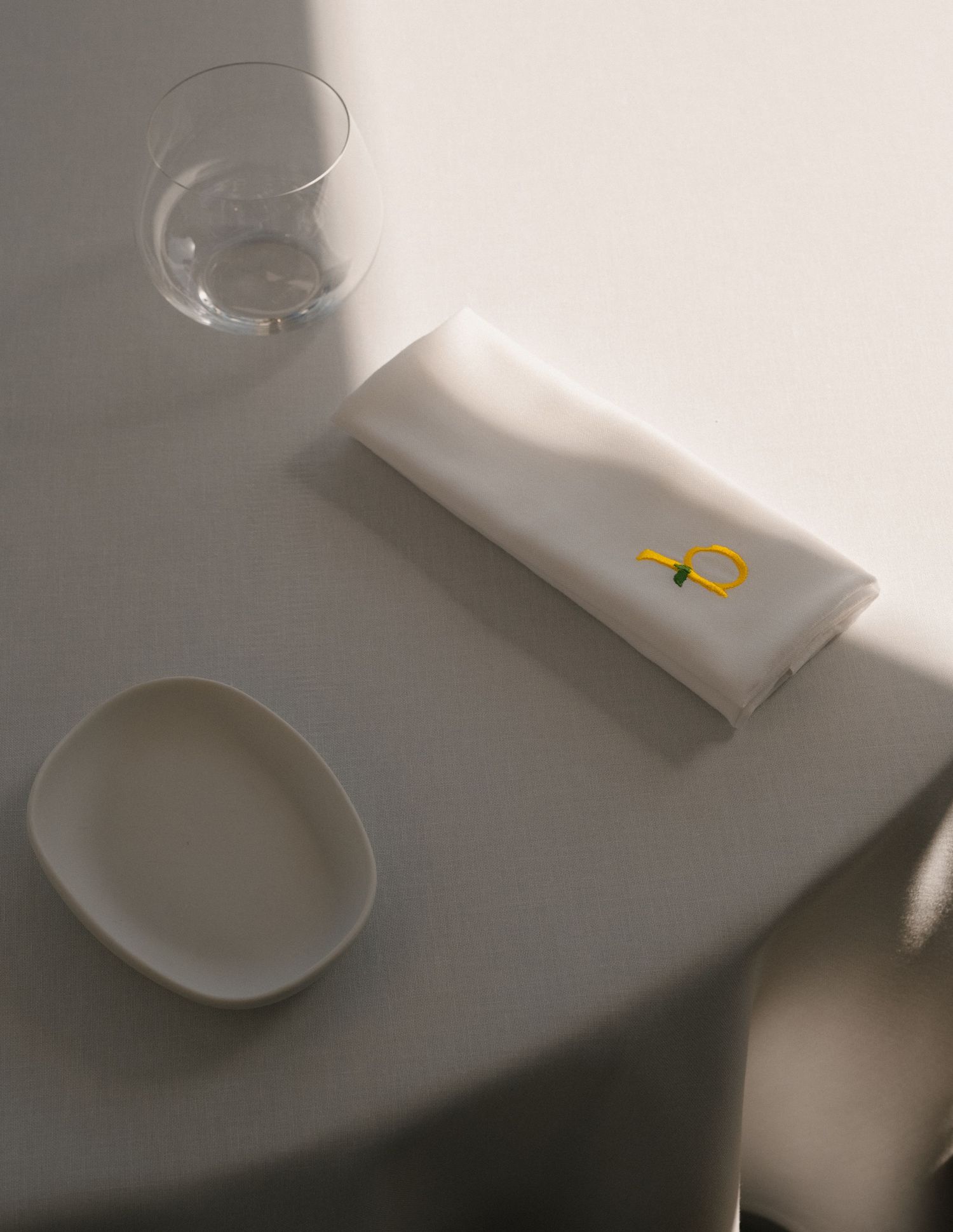 Minimalist table setting with white folded napkin, glass, and ceramic dish on a gray surface.