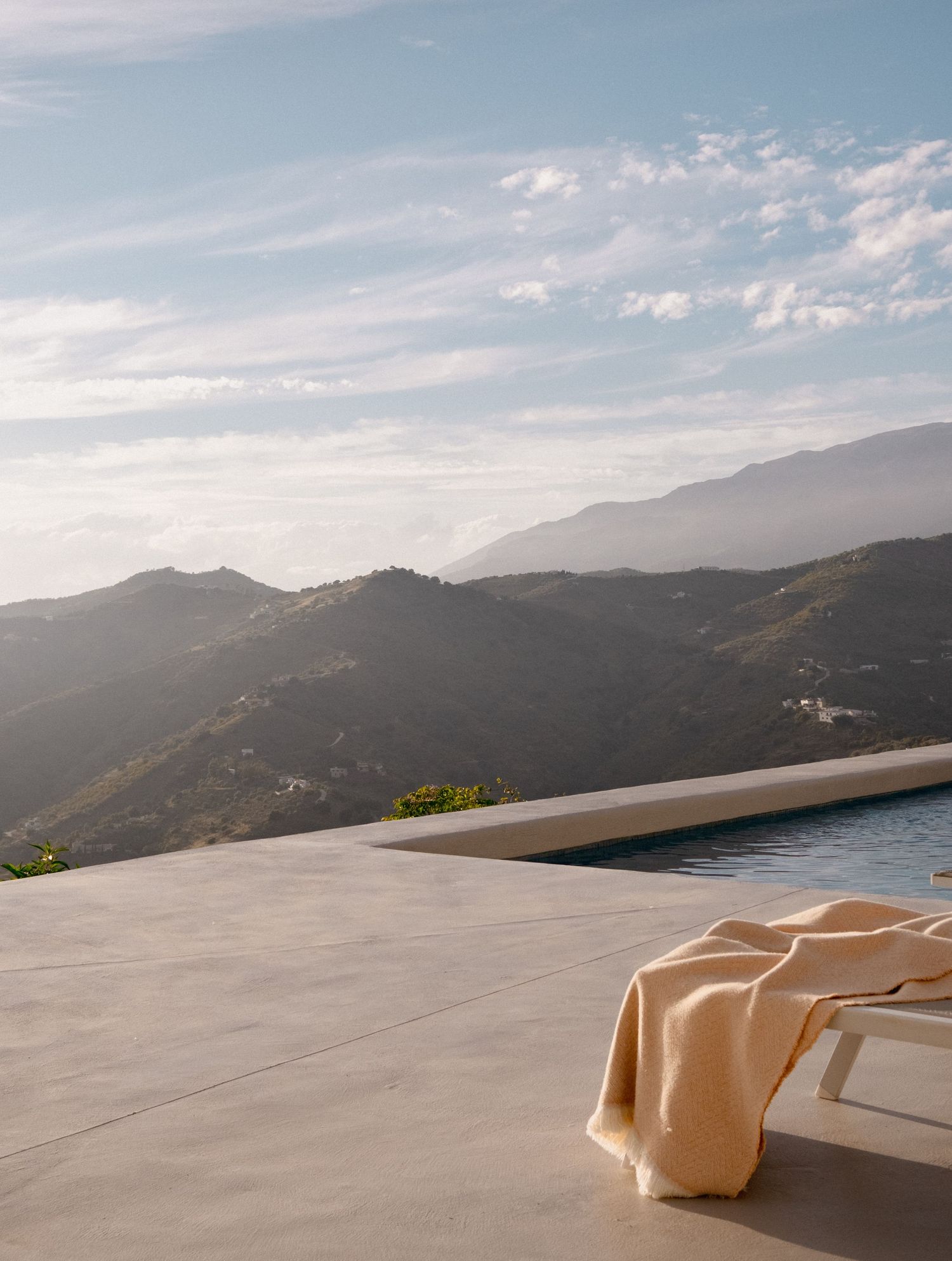 Luxury infinity pool with a cream throw on a lounge chair overlooking misty mountain ranges under a blue sky.
