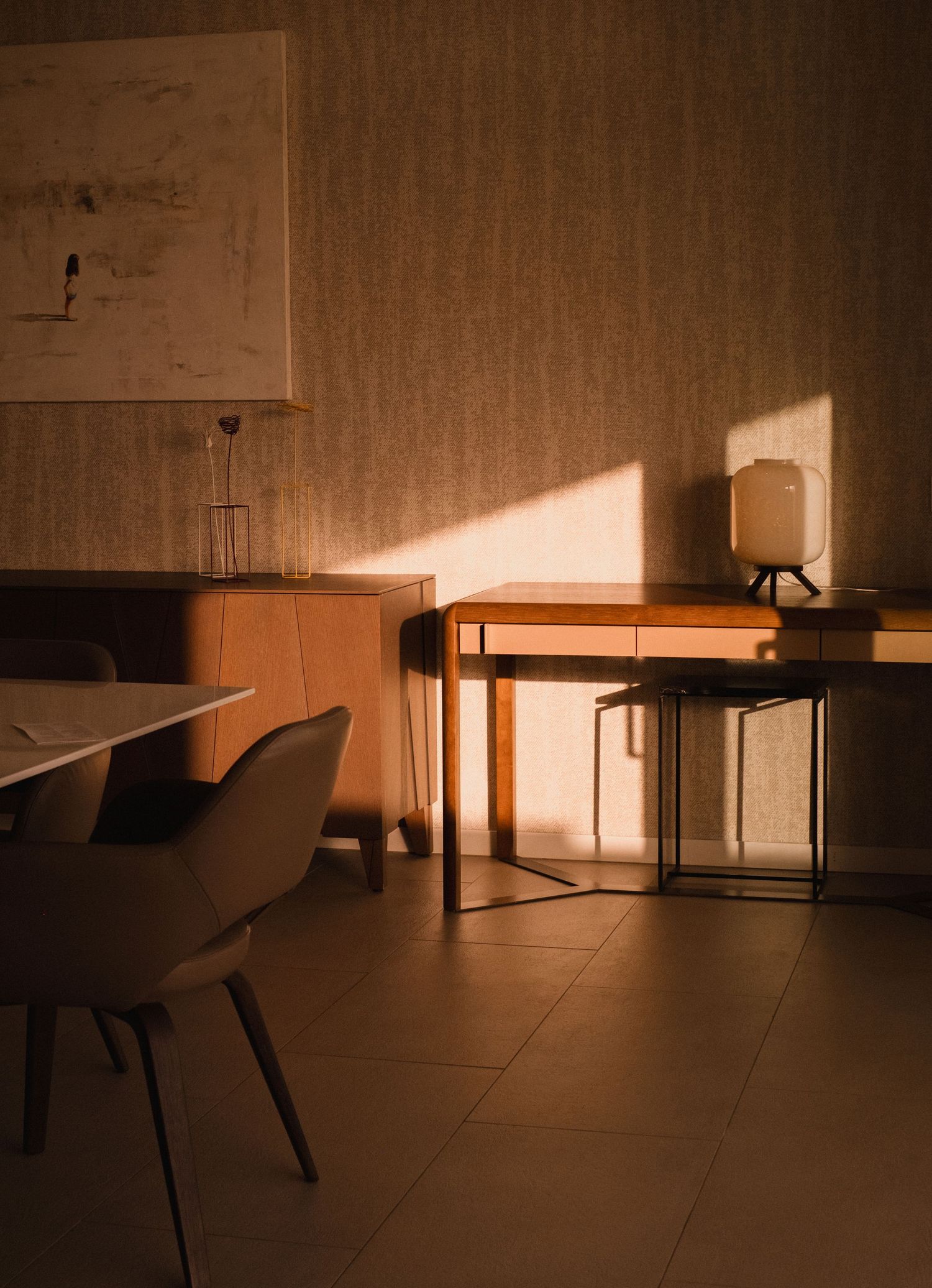 Warm golden sunlight casts shadows across a minimalist room with a wooden desk, chair, and small lamp on tiled floor.
