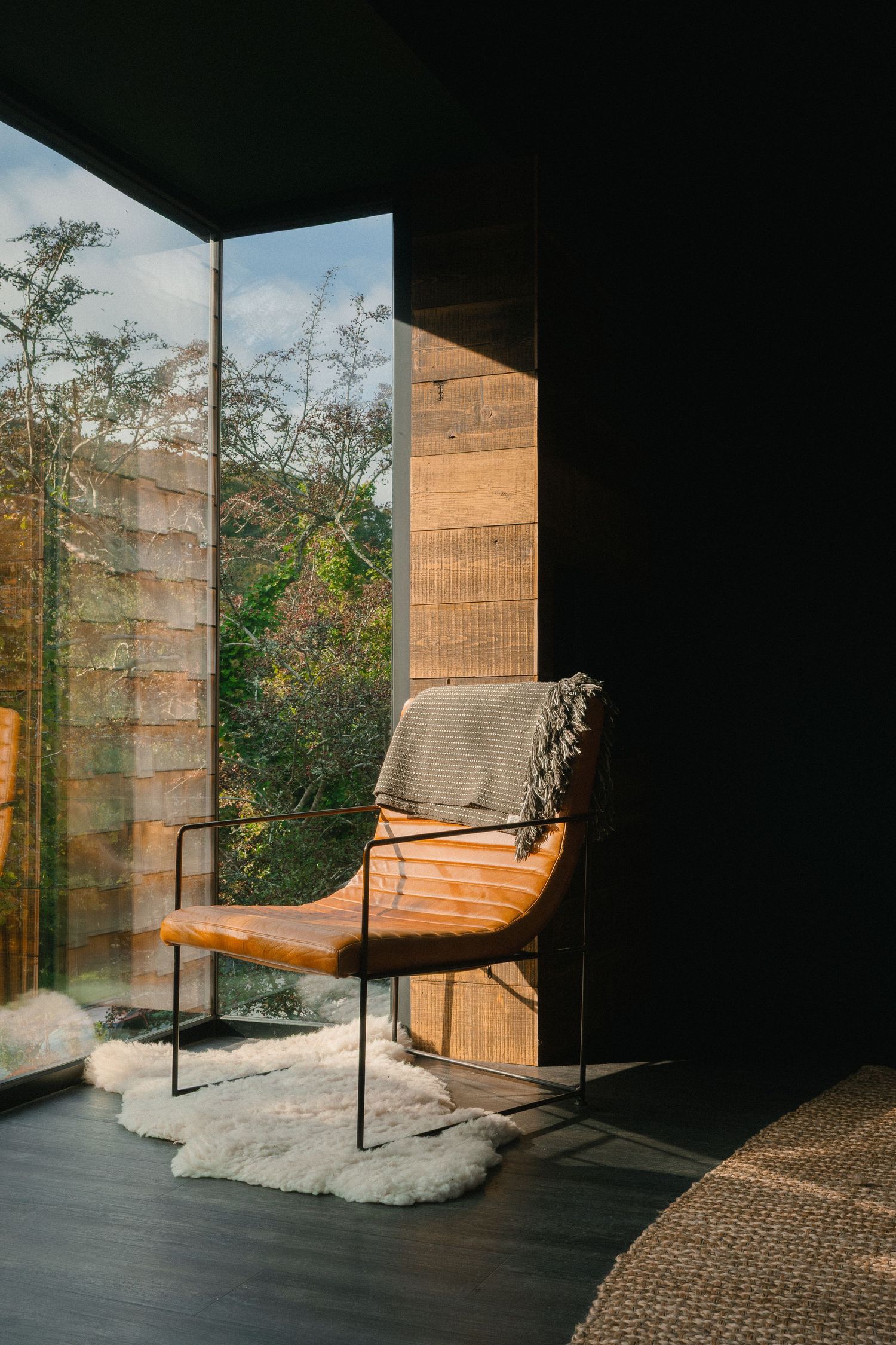 Modern lounge chair sits in sunlit corner with large windows and wooden details.