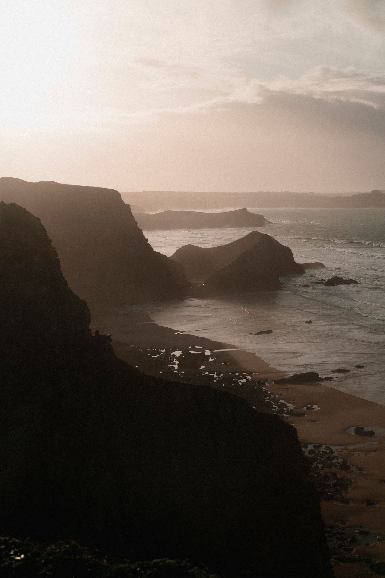 Silhouetted coastal cliffs and rocky formations at dusk with ocean waves crashing below.