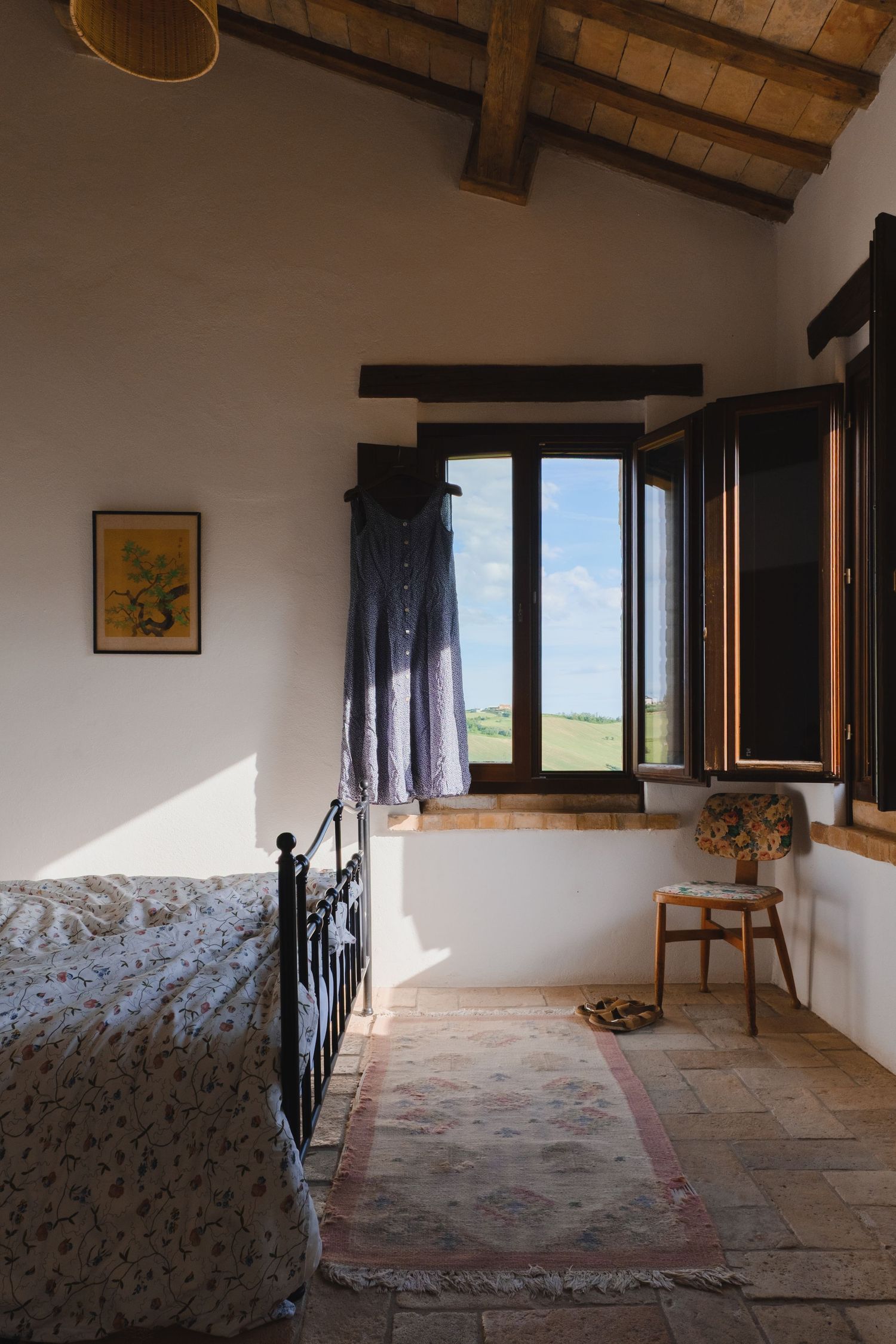 Rustic Tuscan bedroom with wrought iron bed, stone tile floor, wooden beam ceiling, and open shuttered window with countryside view.