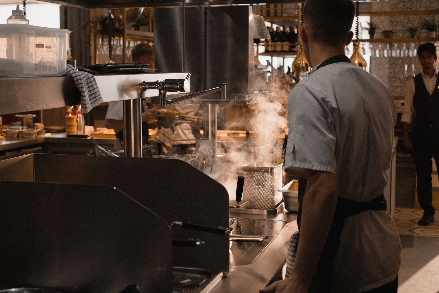 Steam rises from a busy restaurant kitchen as a chef in a gray apron works at a commercial cooking station.