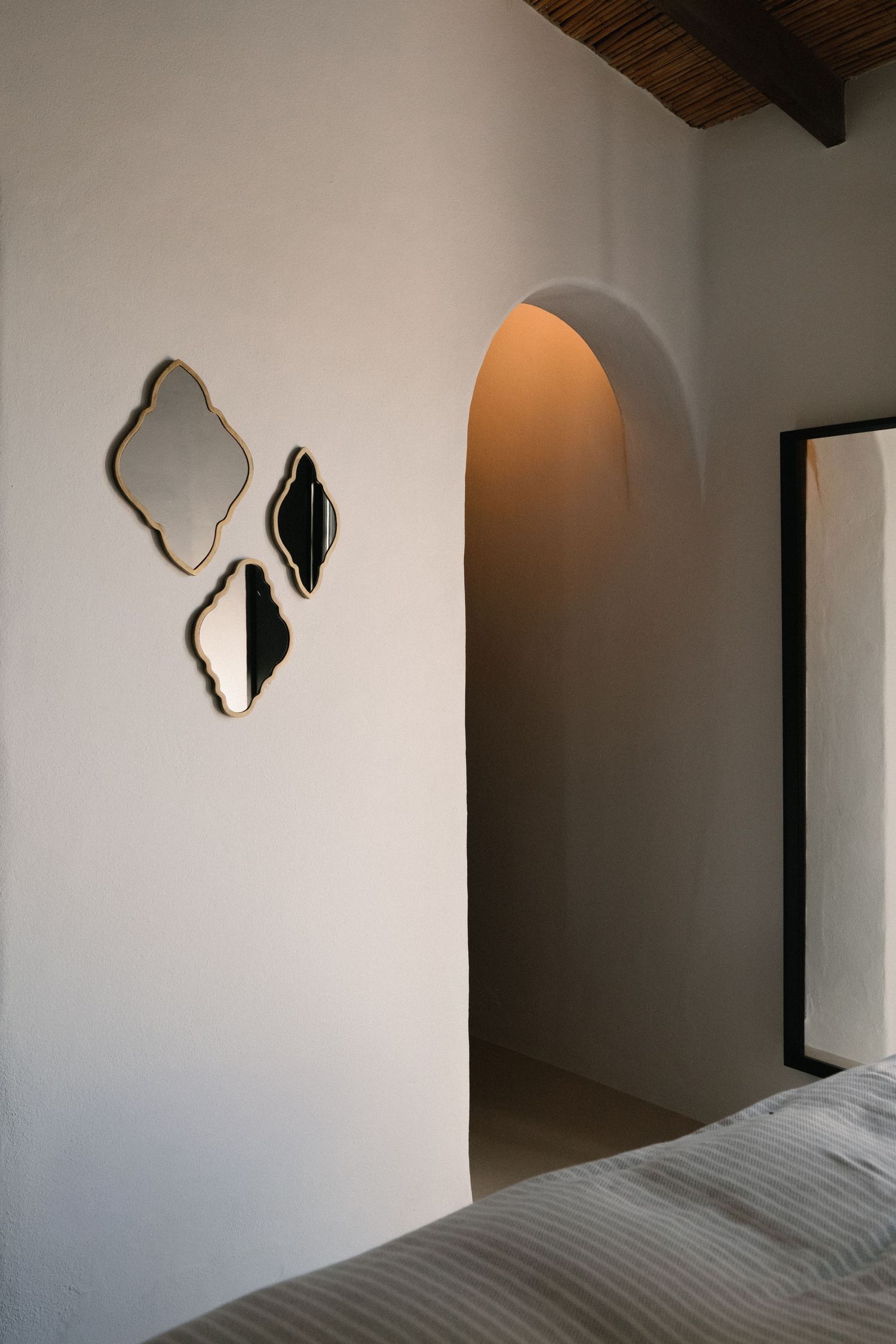 Minimalist bedroom with arched doorway, wooden ceiling, striped bedding, and three decorative gold-framed mirrors on white wall.