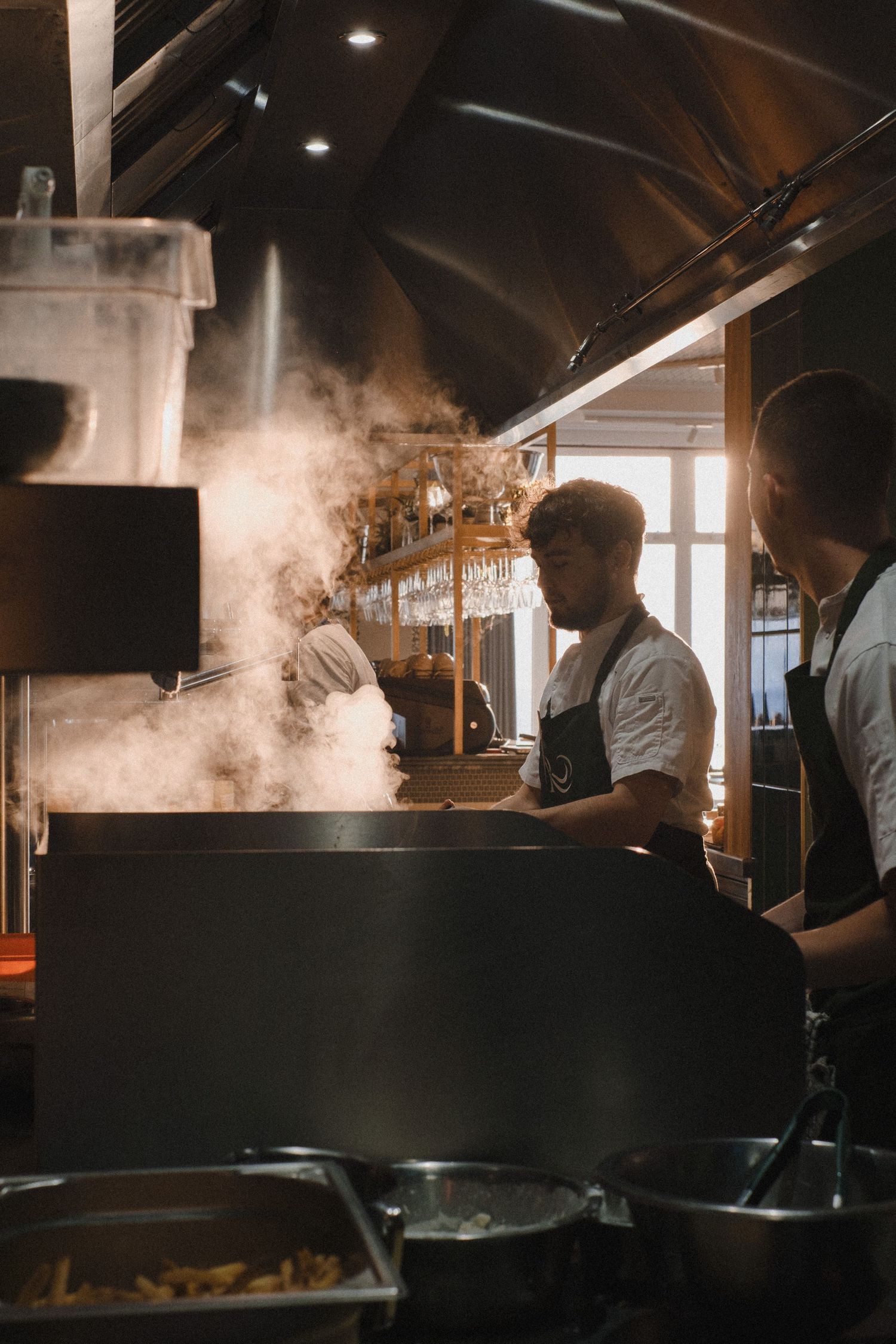 Restaurant kitchen staff working in a dimly lit commercial kitchen with steam rising from cooking stations.