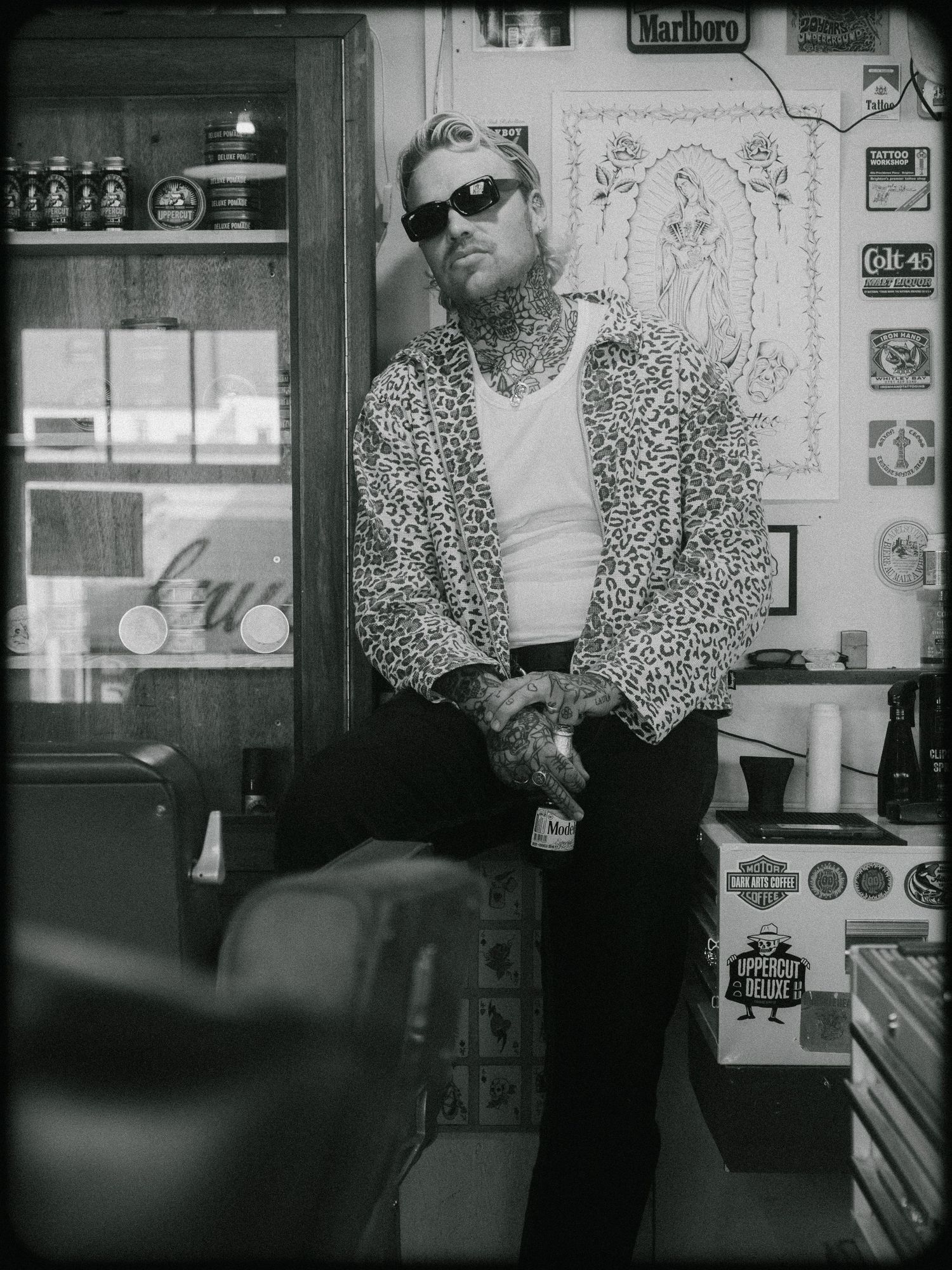 Black and white photo of someone in sunglasses and leopard print jacket leaning against wall in a shop interior.