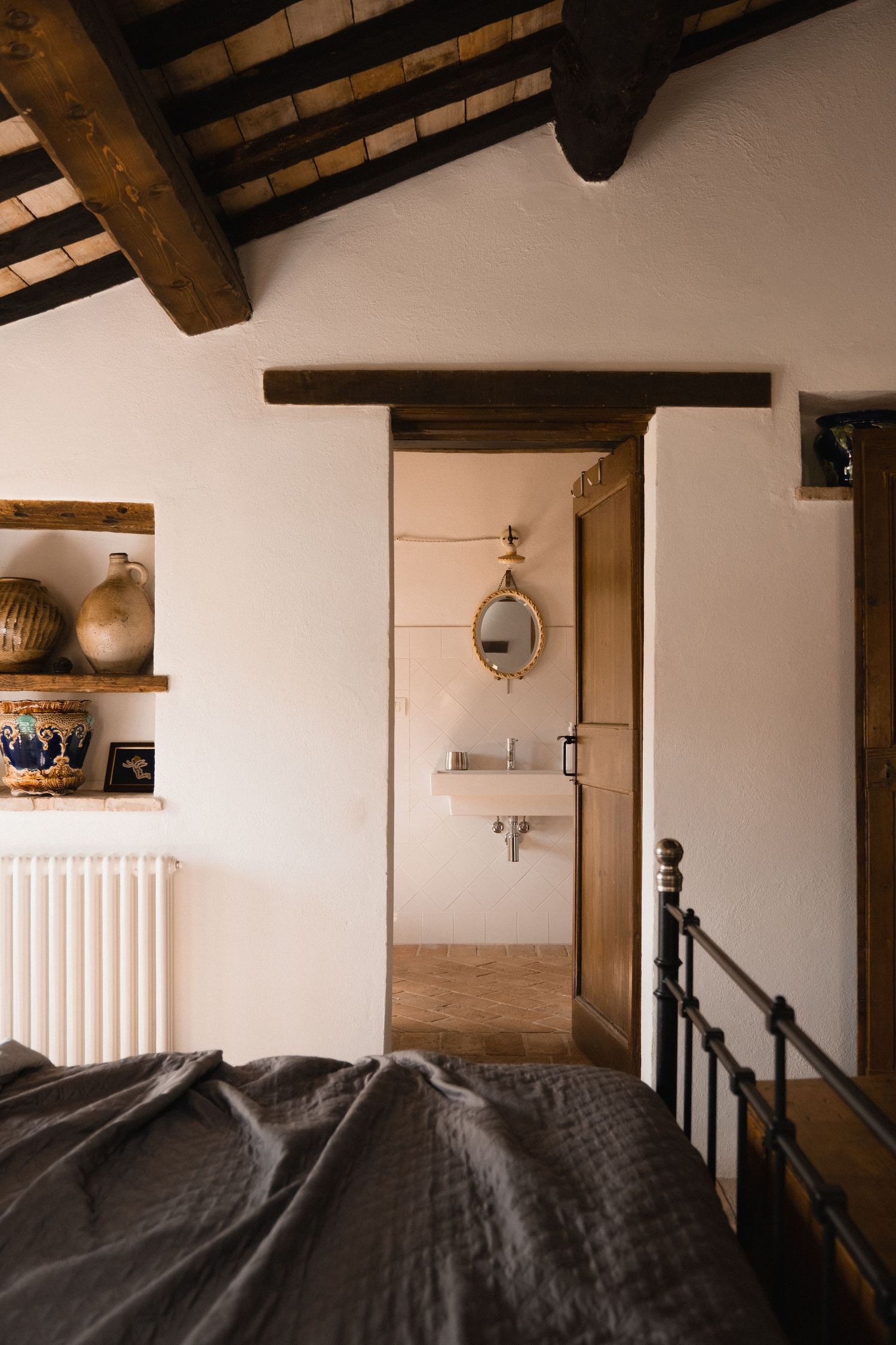 Rustic bedroom with dark linen bedding, wooden beam ceiling, wall shelf with pottery, and en-suite bathroom with oval mirror.