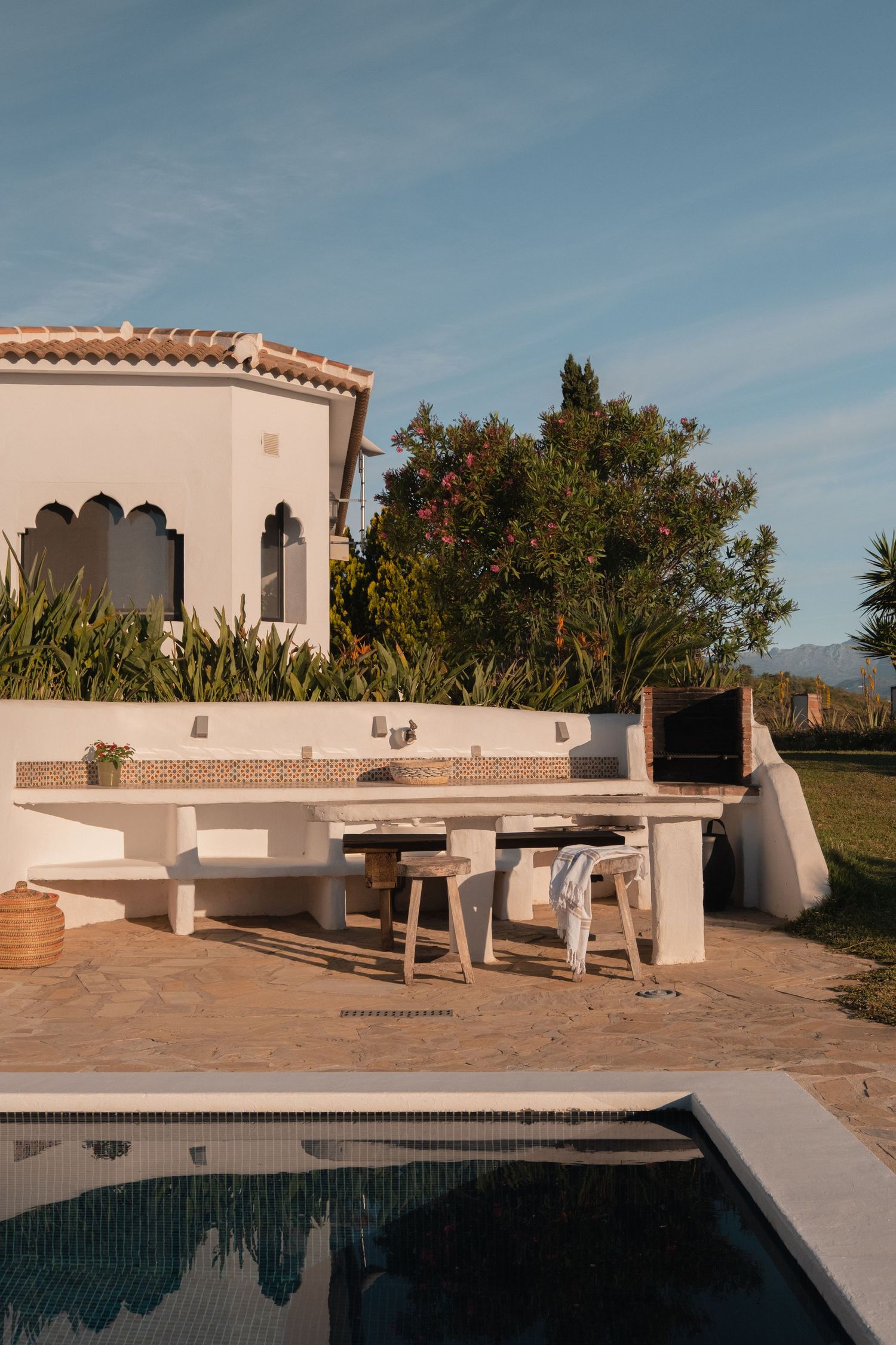 White Mediterranean villa with outdoor kitchen, pool, and lush garden in warm golden light.