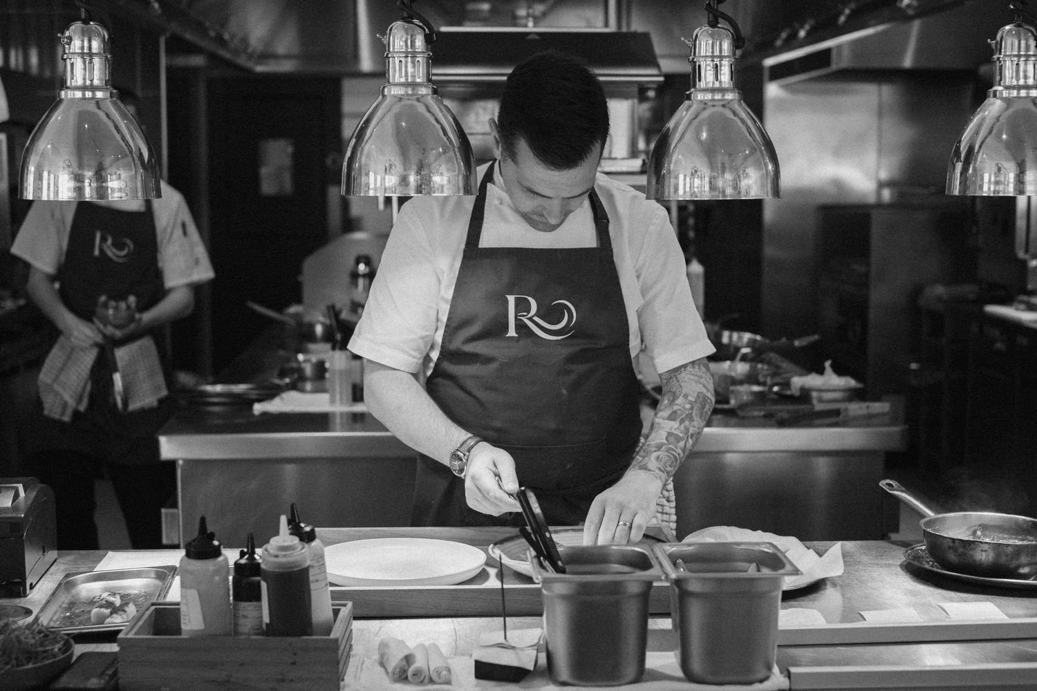 A chef in an apron works at a restaurant kitchen counter with industrial lighting overhead and tattoos visible on their arms.