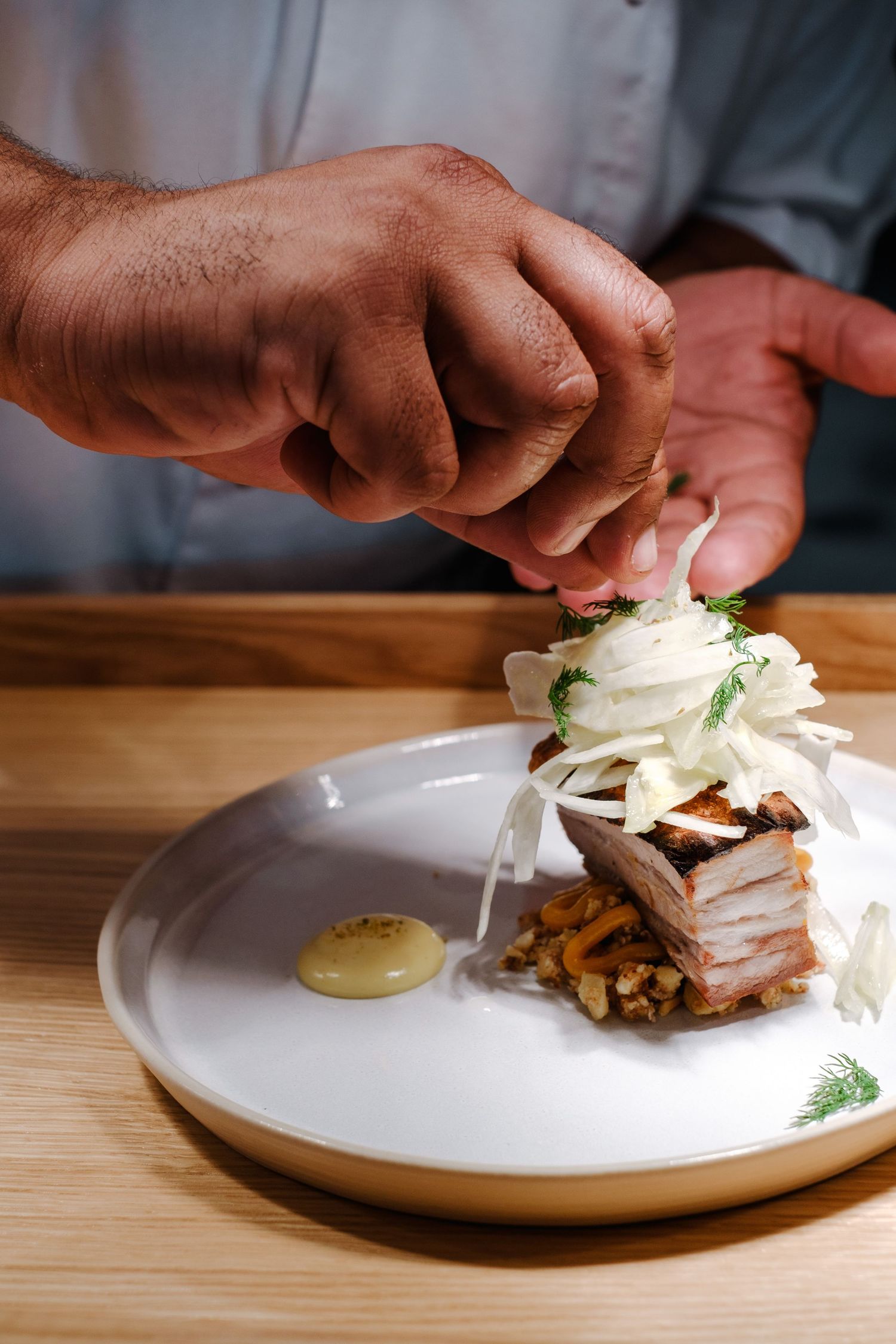Hands assembling a gourmet sandwich with sliced meat and toppings on a white plate in a restaurant kitchen.