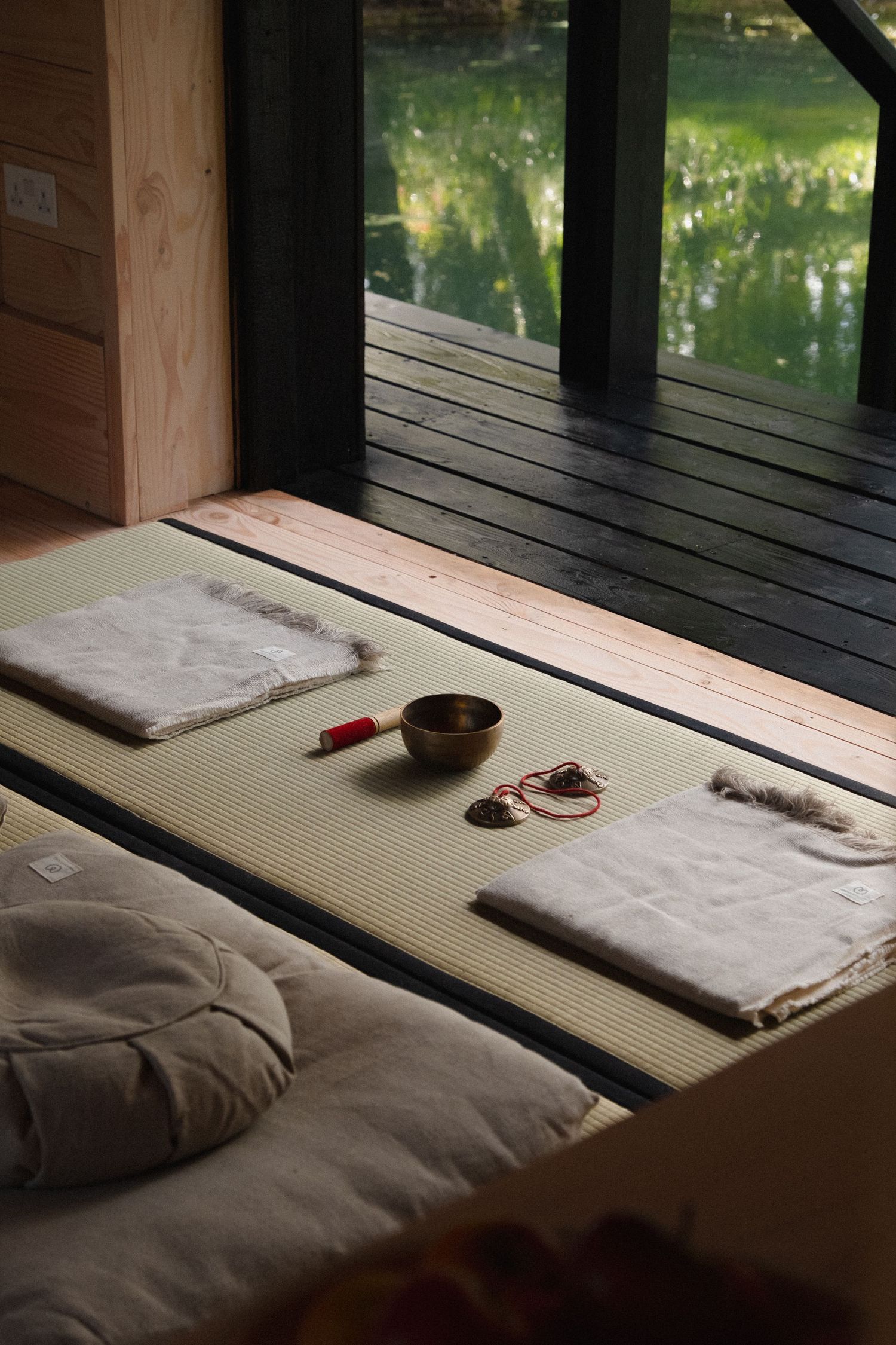 Serene Japanese meditation room with tatami mat, singing bowl, folded linen cushions, and view of tranquil garden pond.